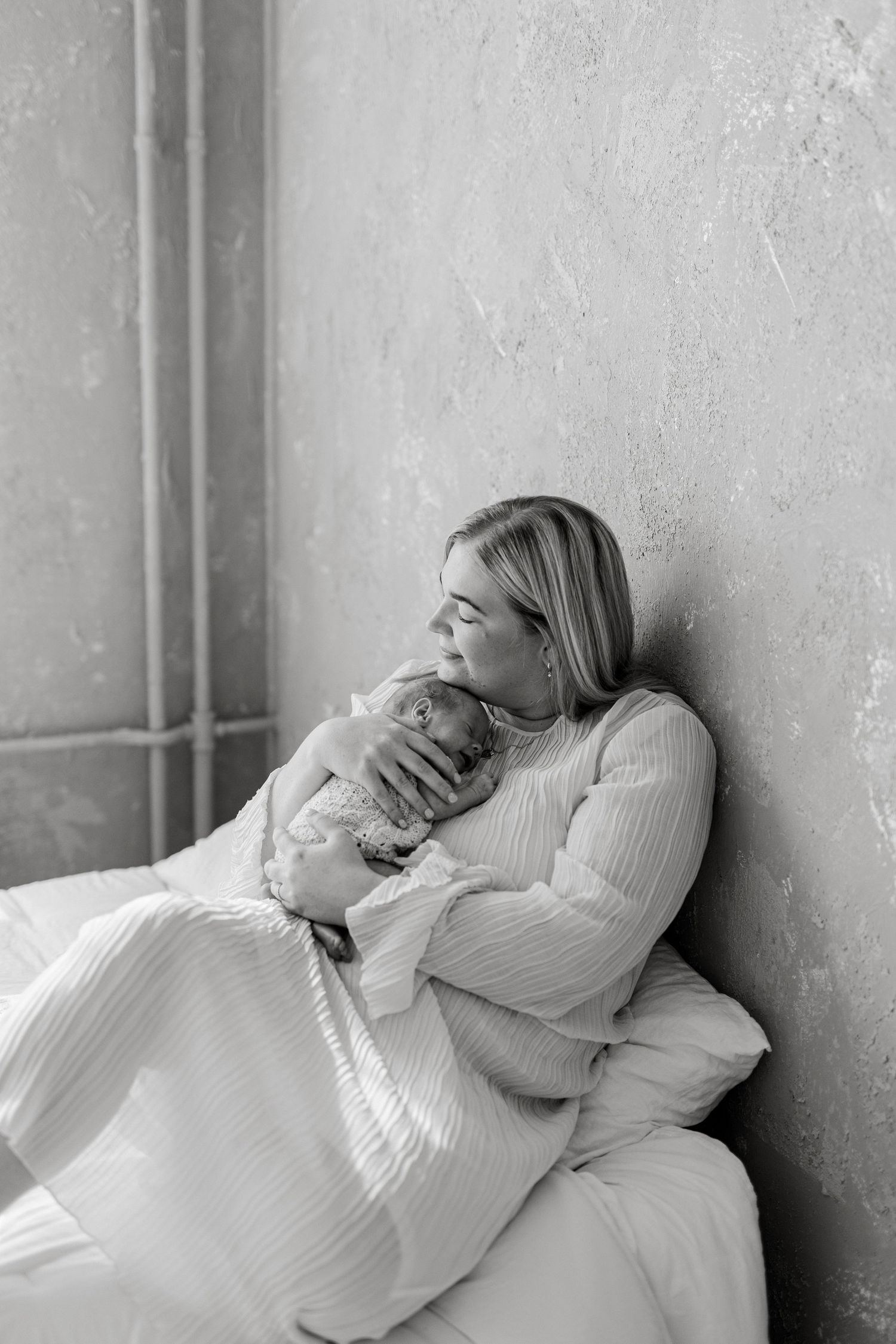 Black and white photo of a mother tenderly kissing her newborn baby while resting against a wall on a bed.