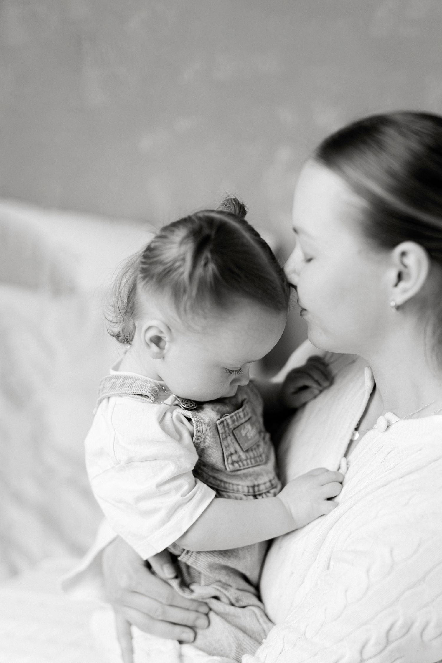 Black and white photo of a young mother kissing her baby while holding the toddler in her arms indoors.