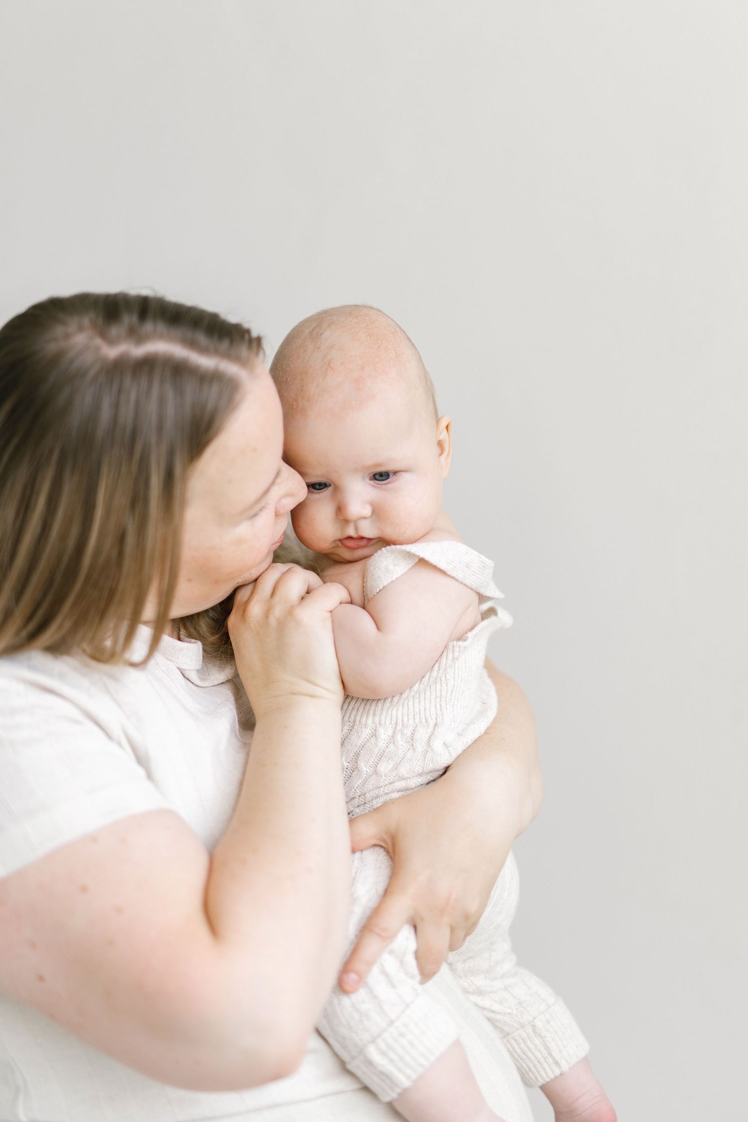 A mother in a white shirt gently kisses her baby dressed in a cream knit romper against a soft white background.