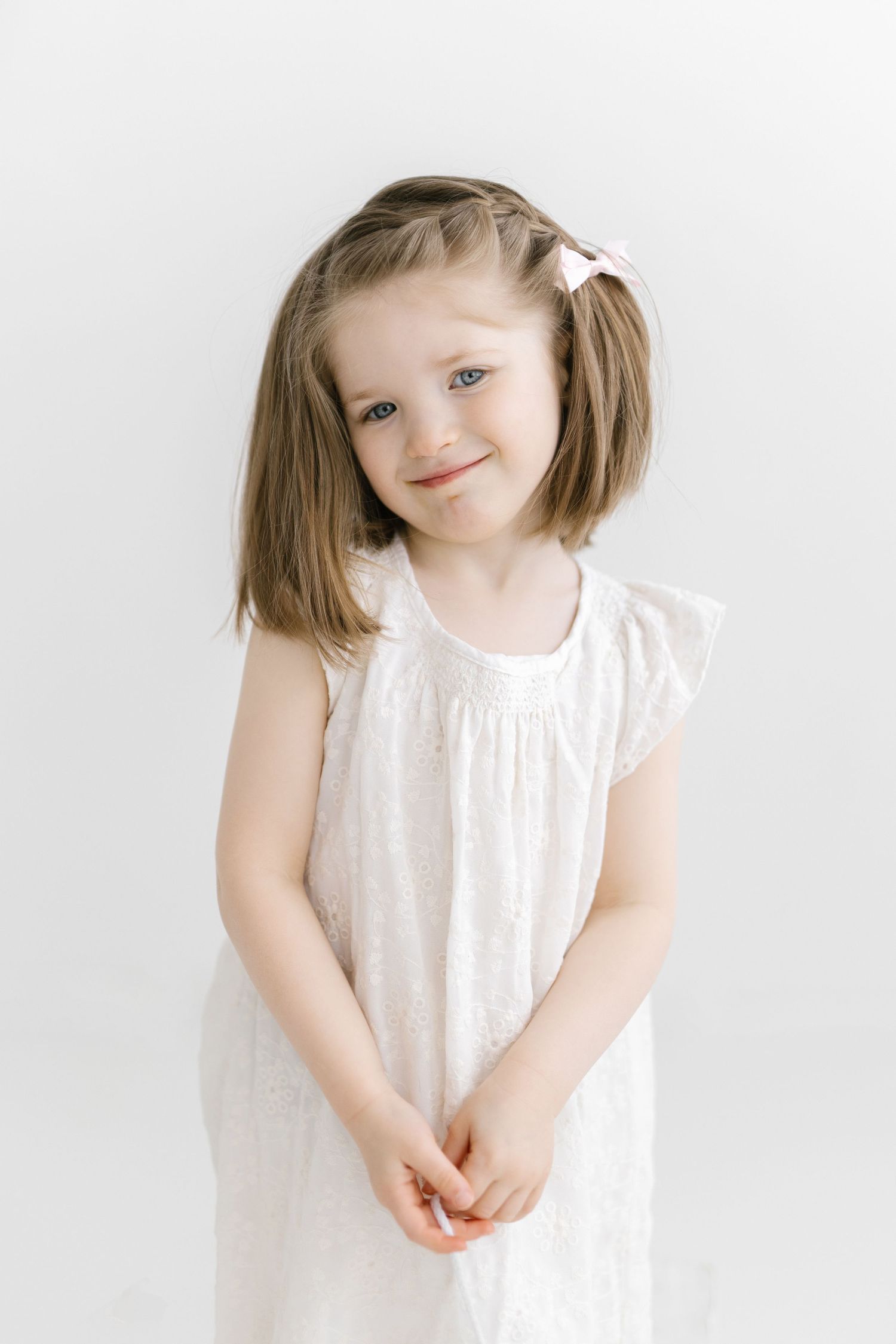 Smiling young girl in white dress with pink hair bow posing against white background.