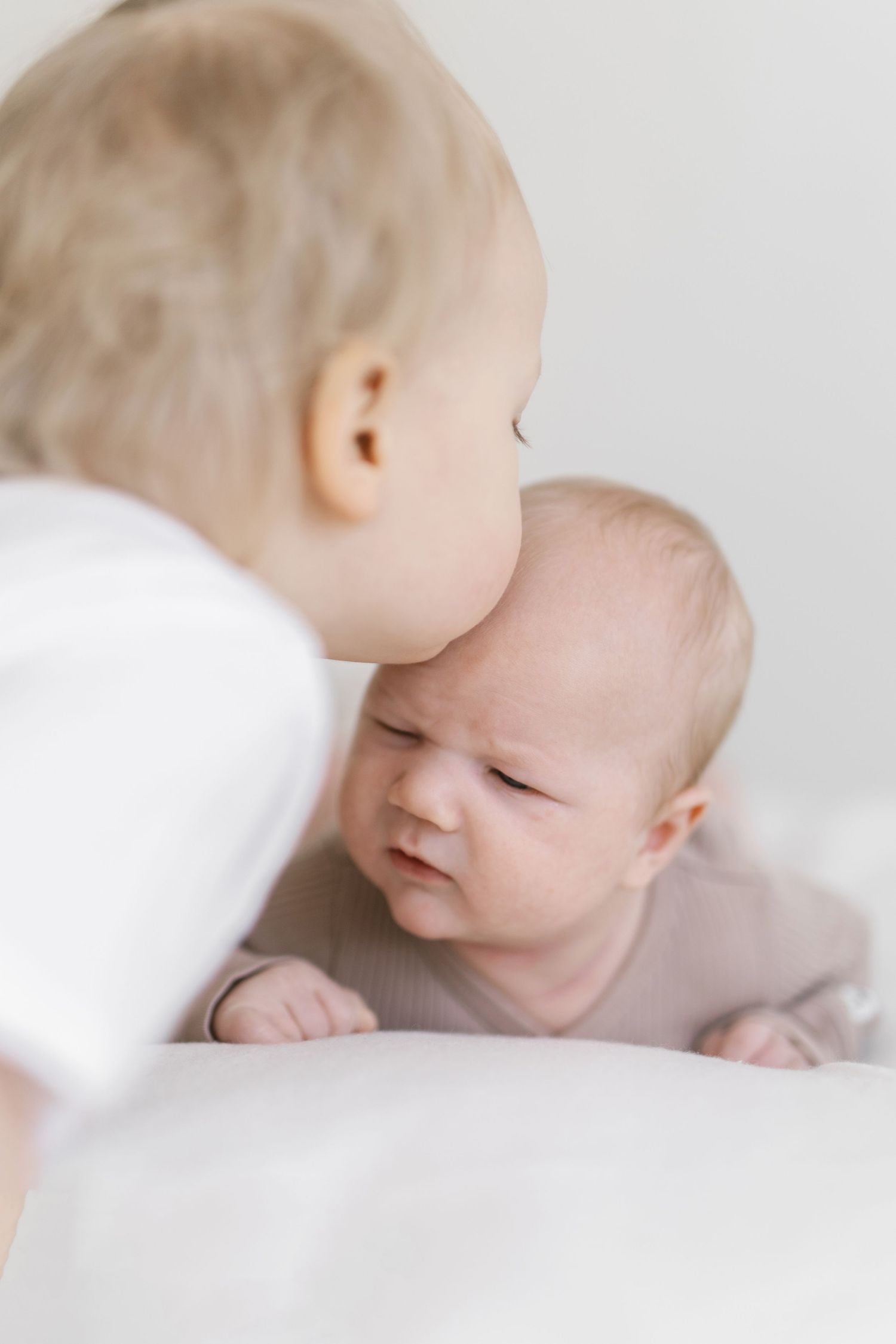 Two babies interact closely, one kissing the other on the cheek while the younger infant looks up with a curious expression.