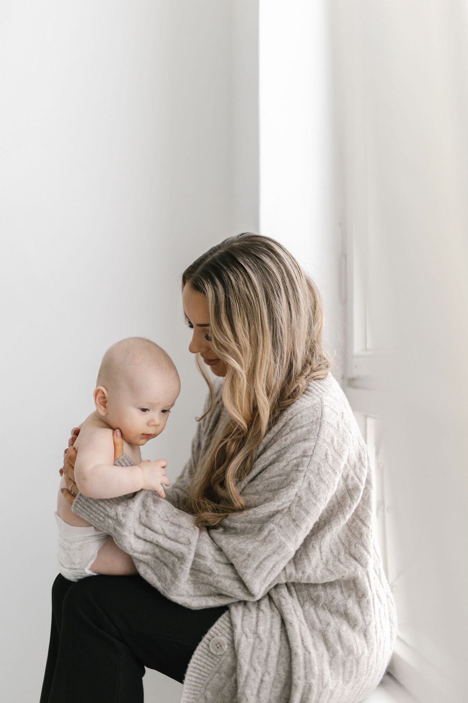 A mother with long blonde hair holds her baby near a bright window, wearing a cozy knit cardigan.