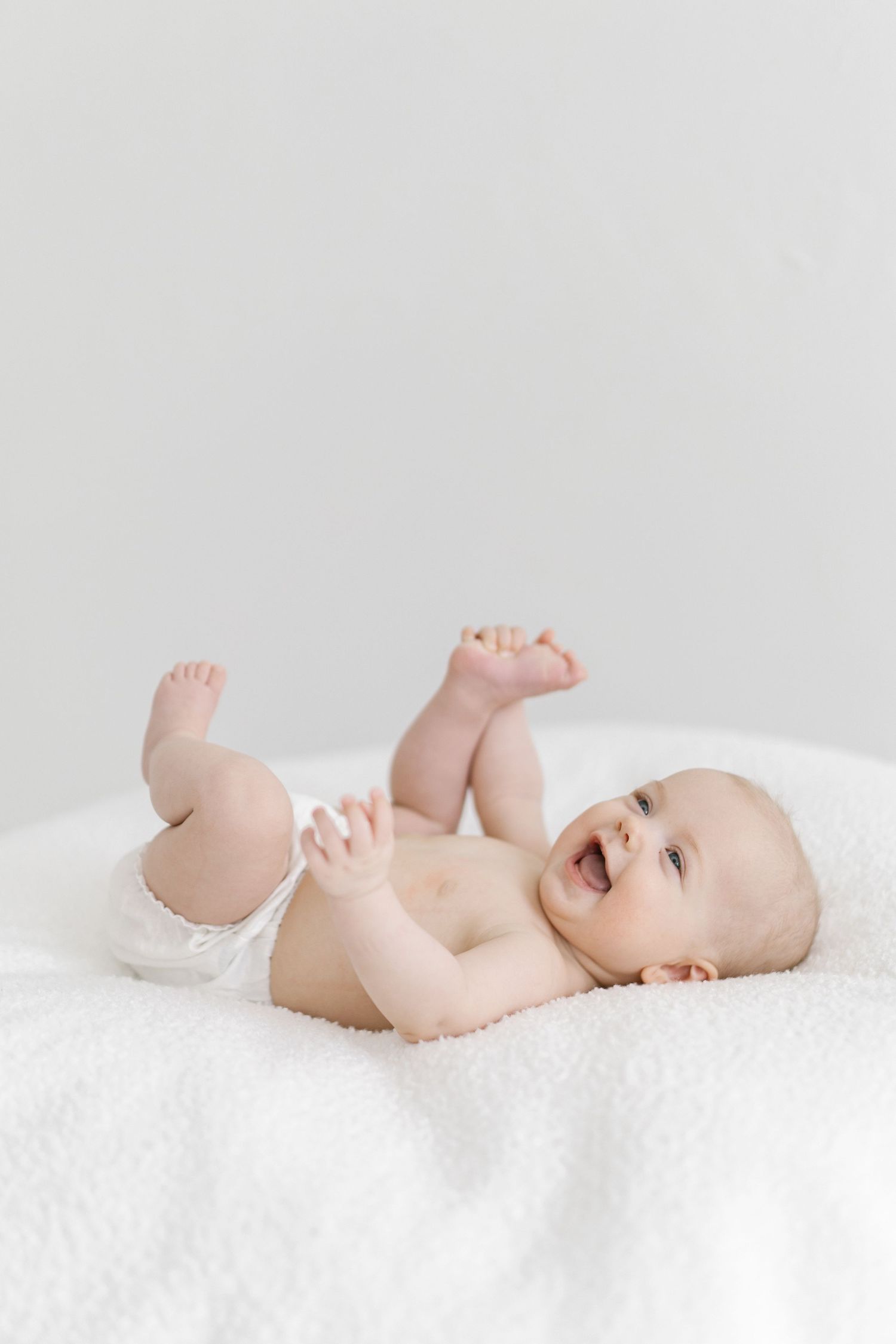 Happy baby lying on white blanket, laughing and playing with feet, wearing diaper in bright minimalist setting.