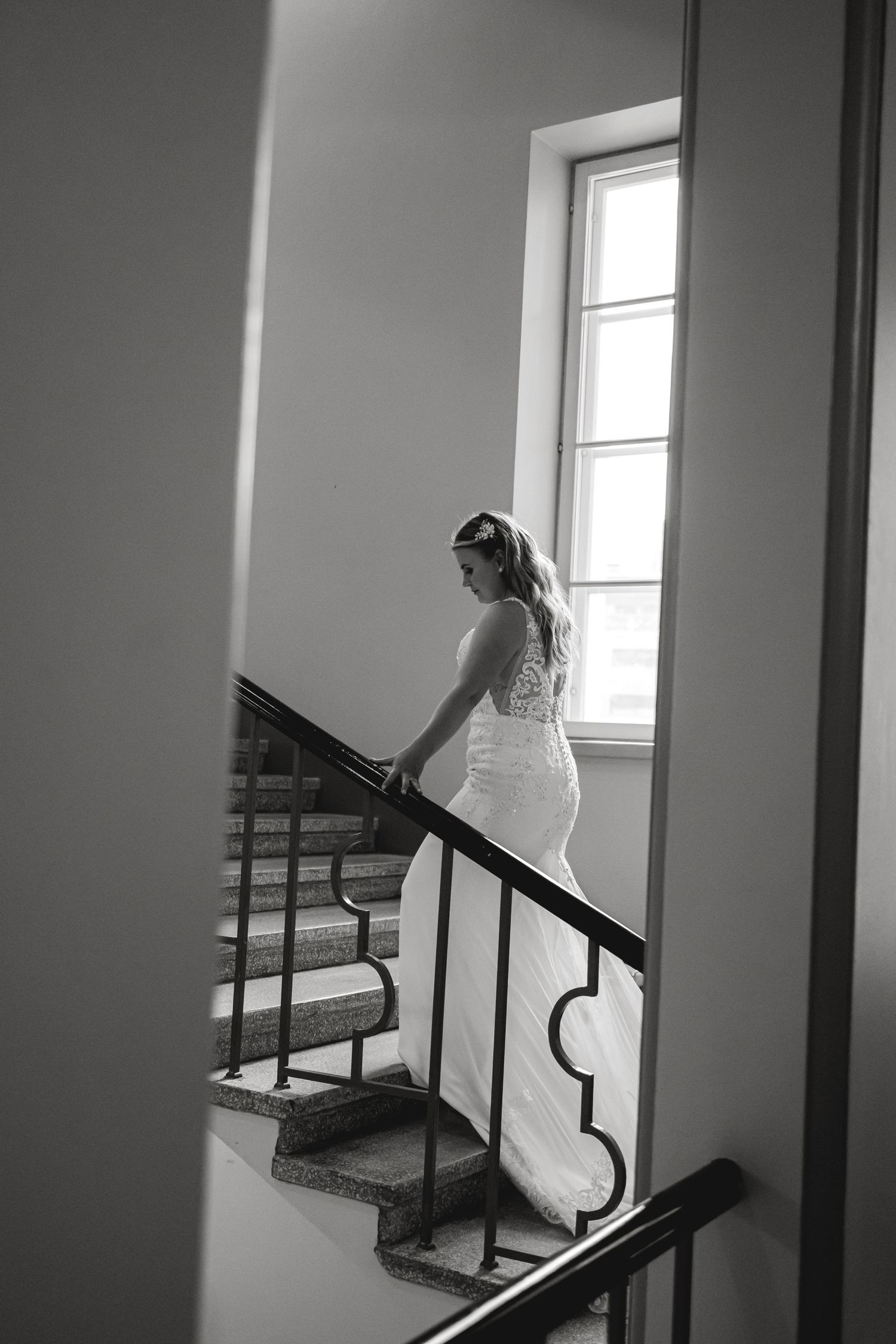 A bride in a fitted lace gown ascends a staircase near a bright window.
