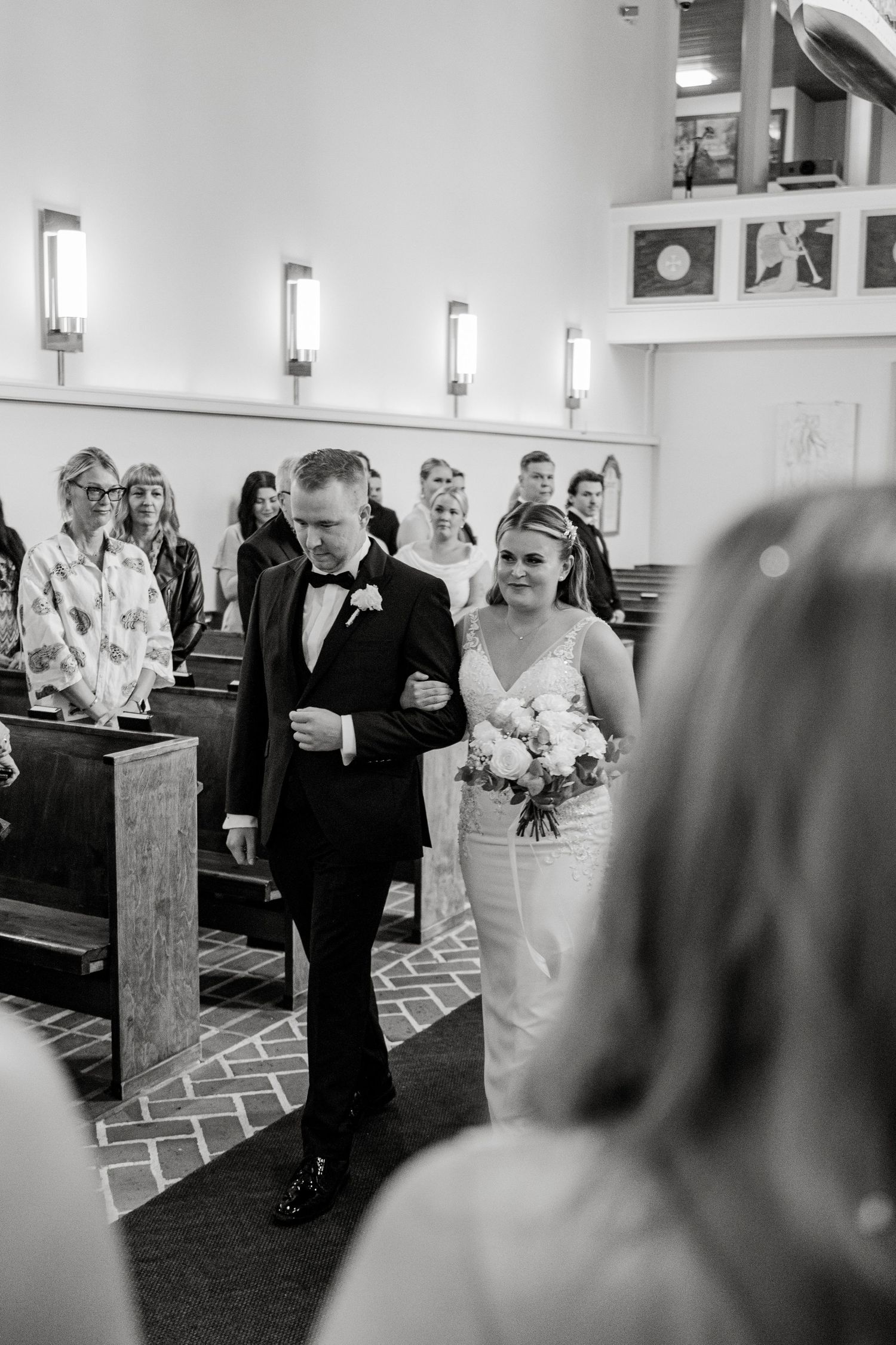 Black and white wedding ceremony processional in a traditional church with guests seated in pews.