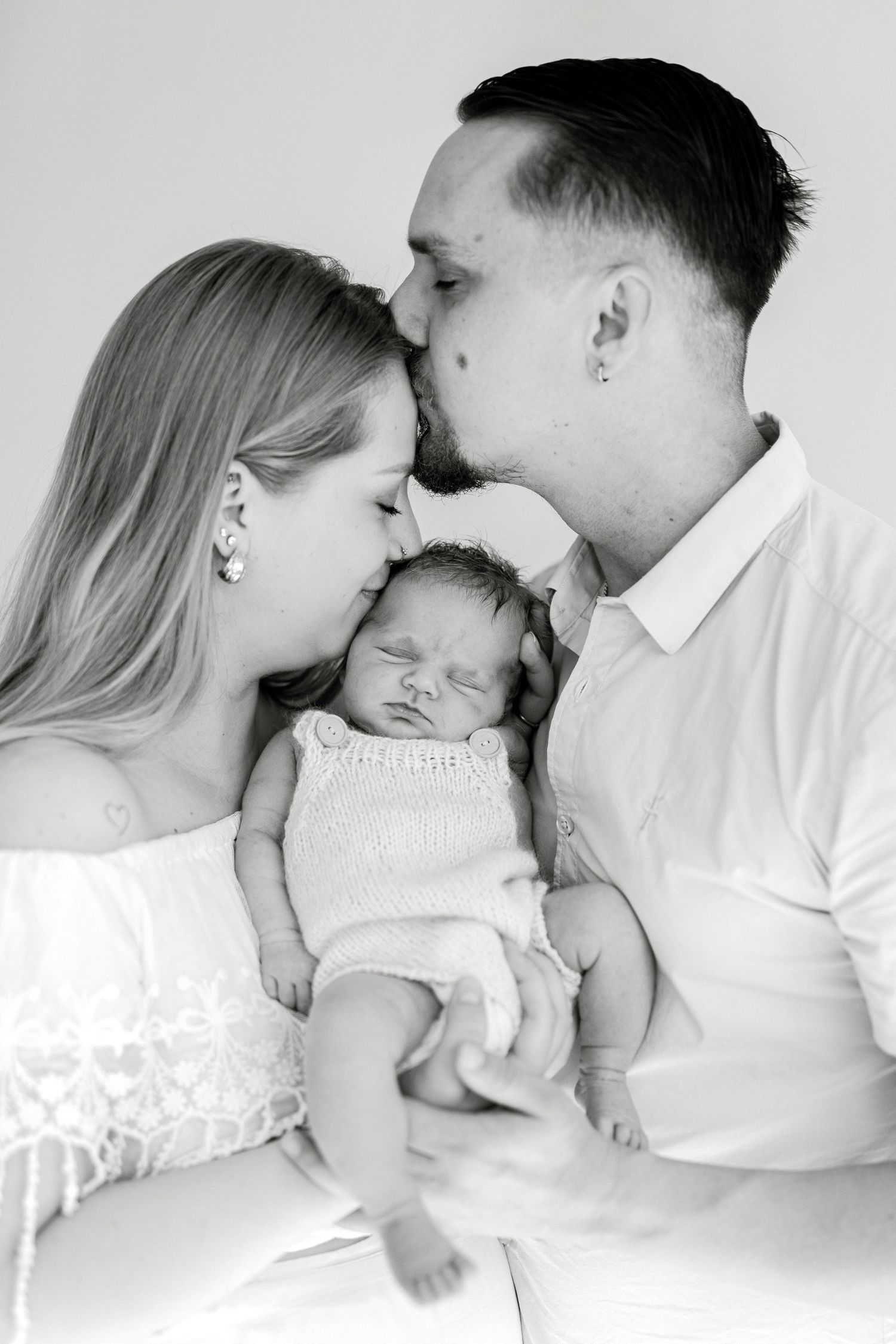 Black and white photo of parents kissing their sleeping newborn baby, a tender family portrait moment.