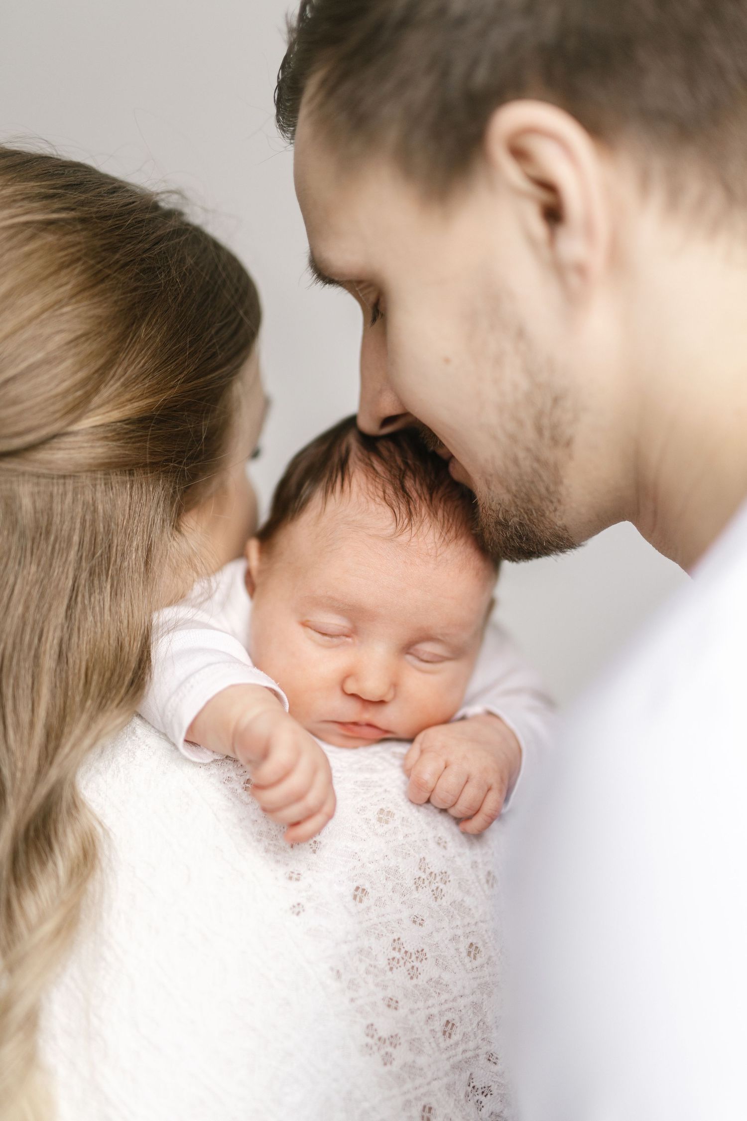 Parents tenderly kissing their sleeping newborn baby wrapped in white swaddle blanket.