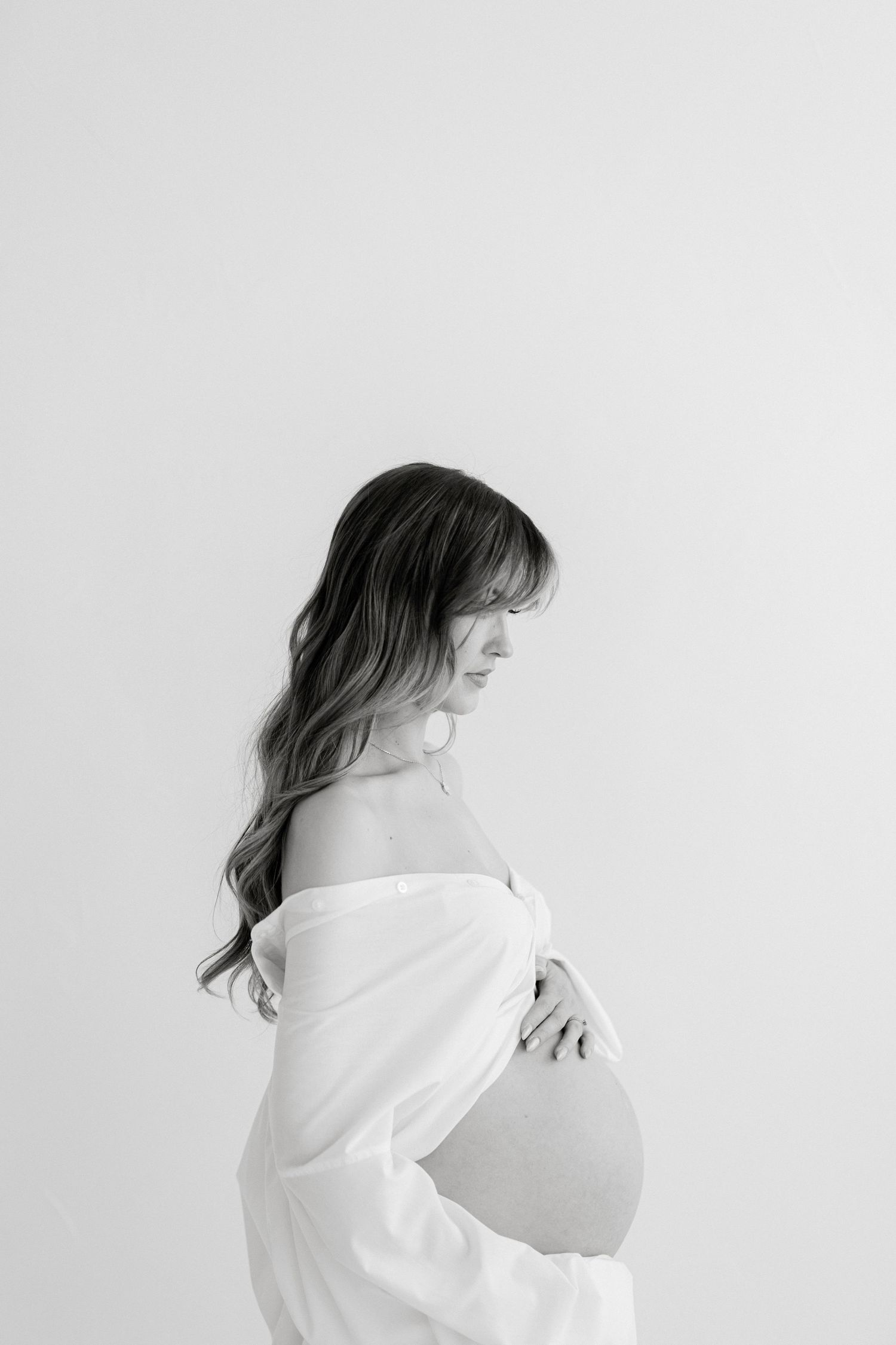 Black and white maternity photo of a woman with long hair cradling her baby bump in a white off-shoulder top.