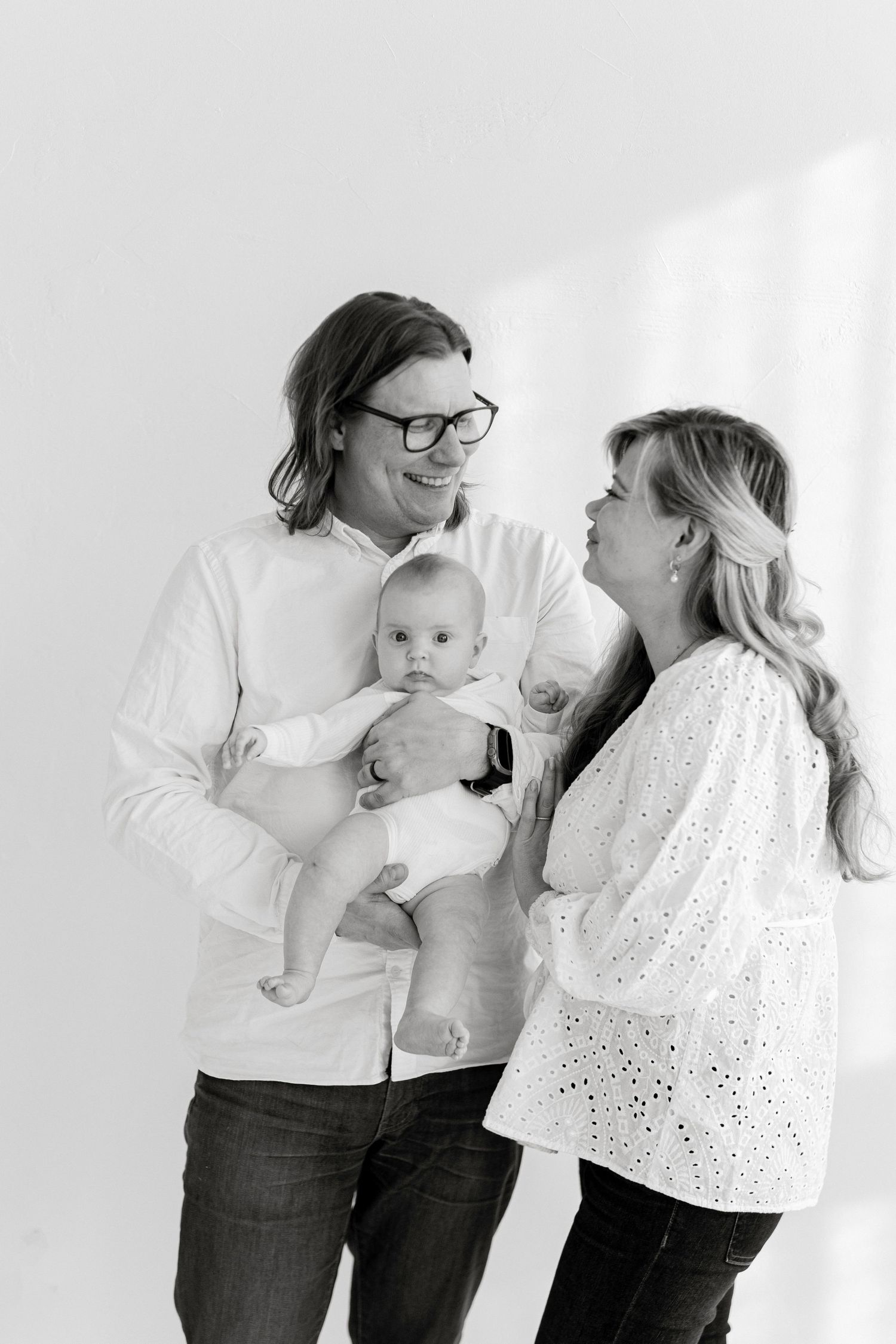 Black and white photo of smiling parents holding their newborn baby in a bright studio setting.
