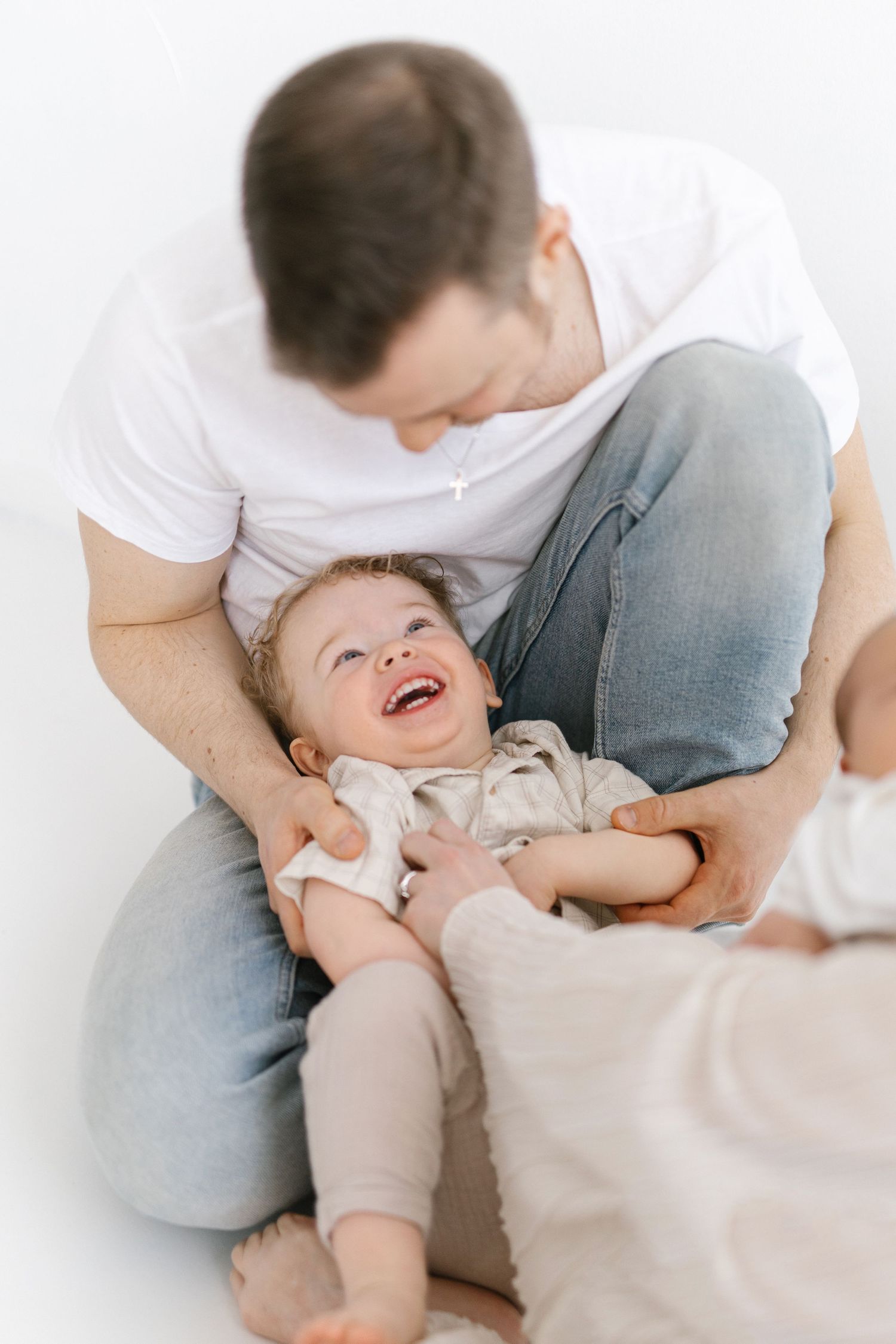A laughing baby rests in a father's lap during a joyful family photo session on a white background.