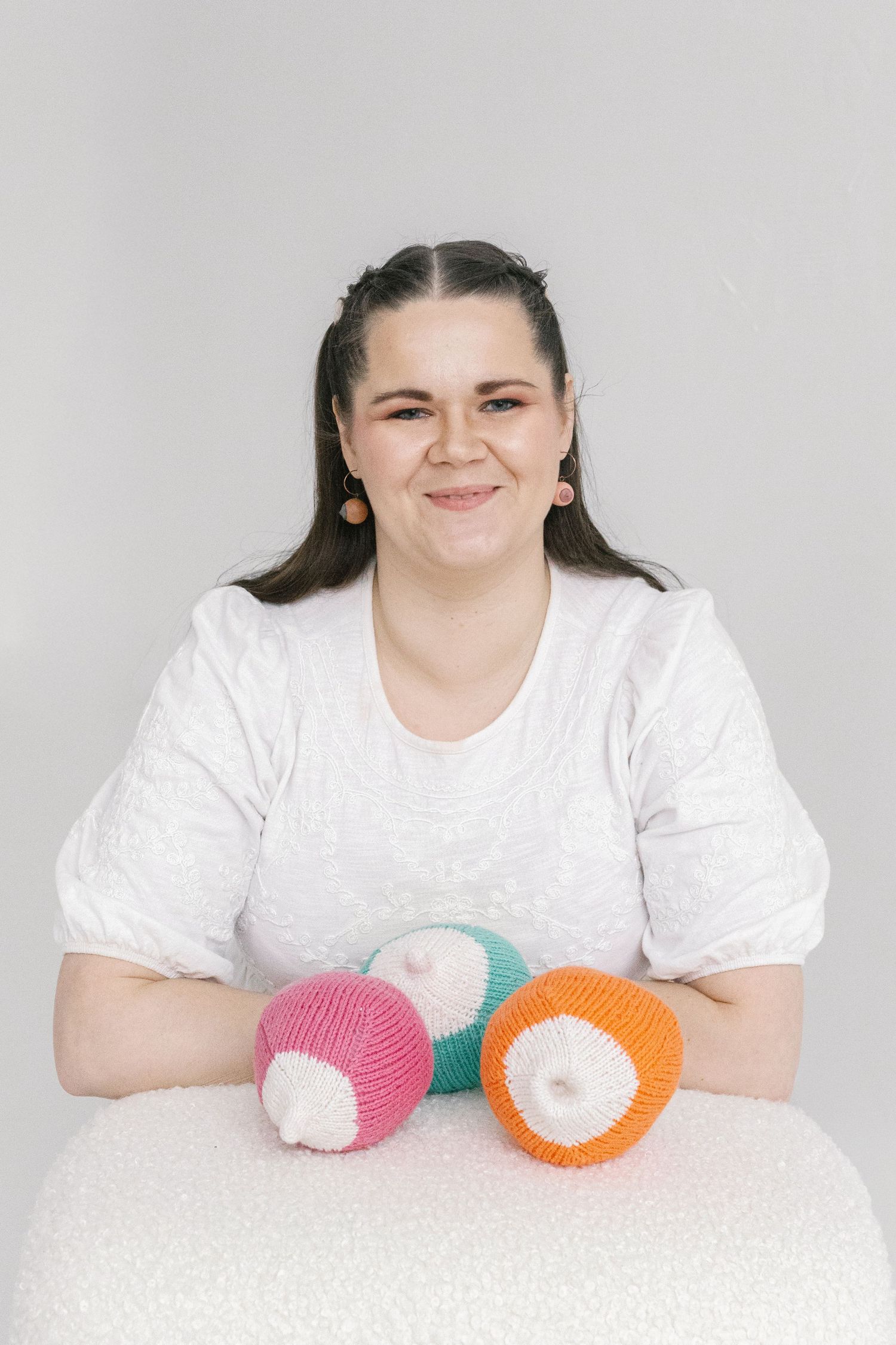 Smiling woman in white top poses with three colorful knitted balls on a white surface.