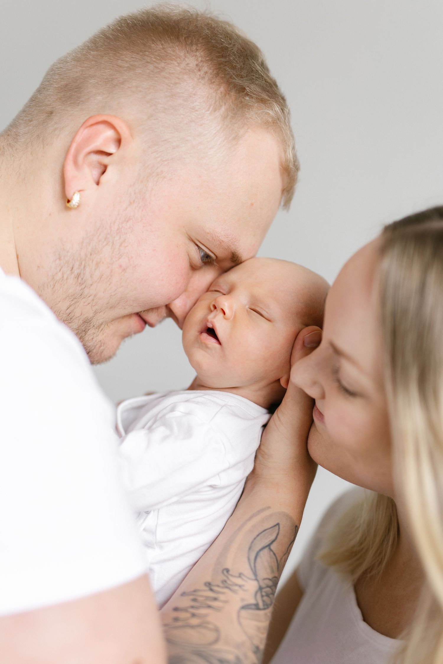 Parents in white shirts lovingly kiss their smiling newborn baby, creating a tender family portrait.