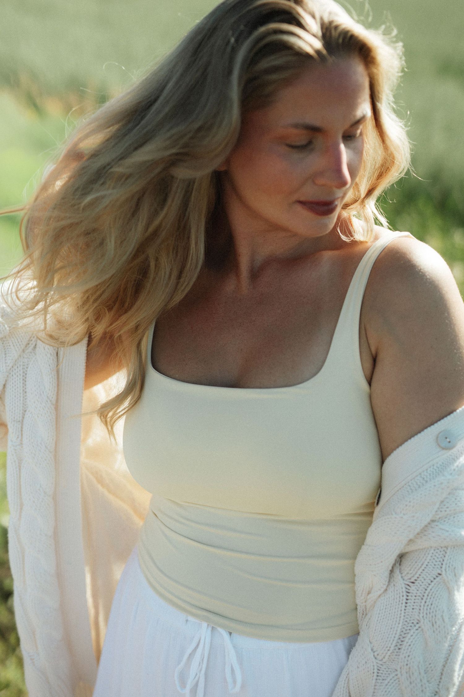 Blonde woman in cream tank top and white cardigan posing outdoors in natural sunlight.