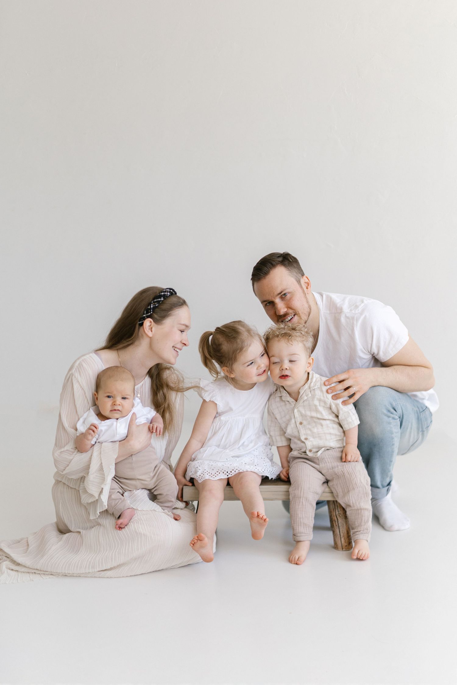 A young family with three small children poses together on a wooden bench against a white studio background.