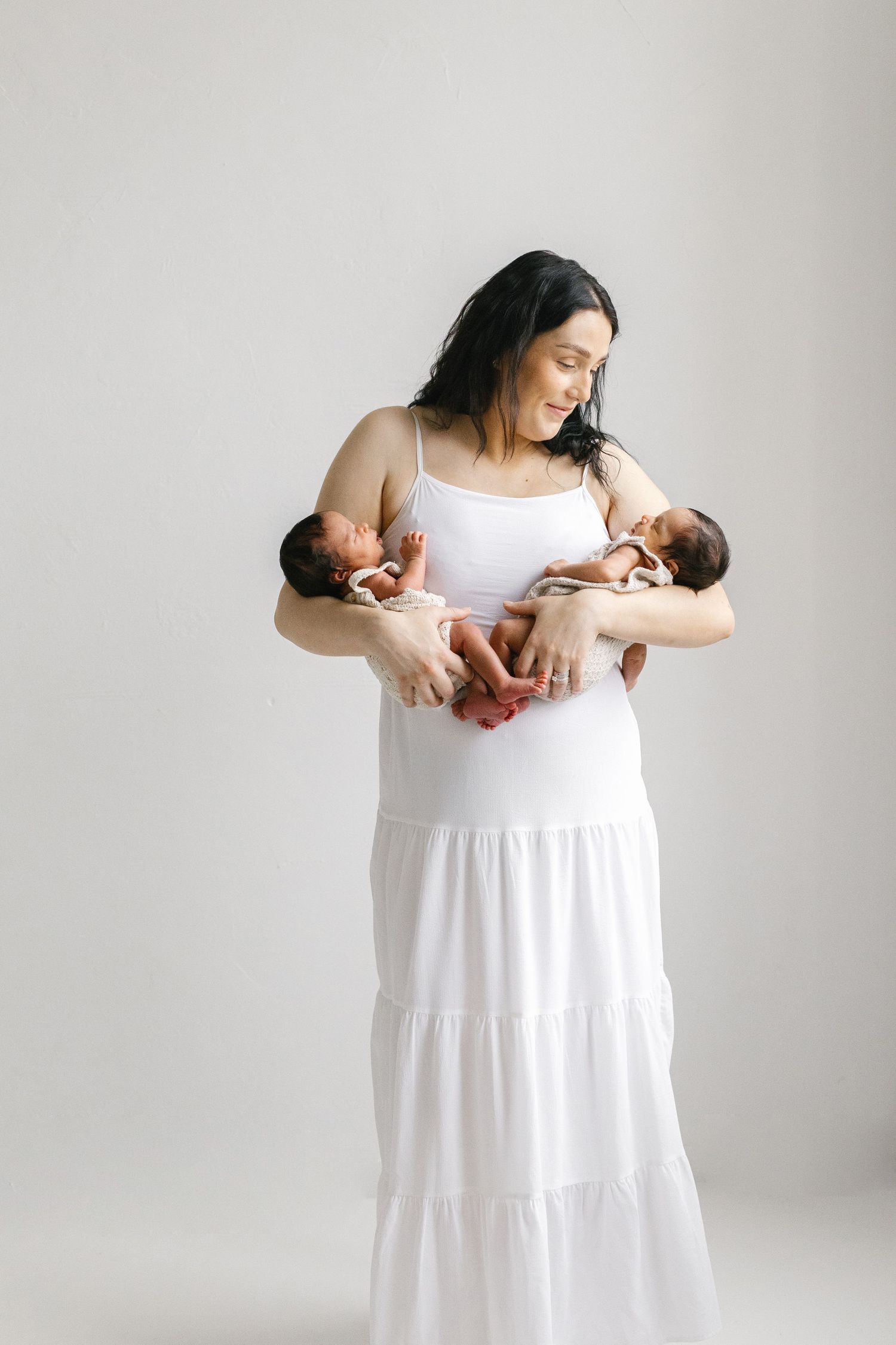 A mother in a white dress holds her newborn twins, smiling warmly in a bright studio setting.
