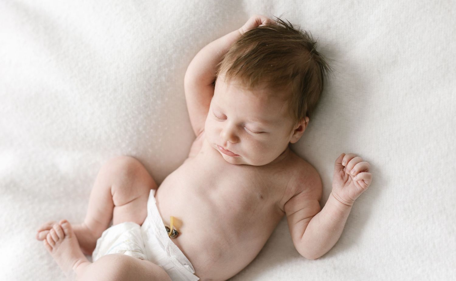 Sleeping newborn baby in a white diaper lying on a soft white blanket with one arm raised above the head.