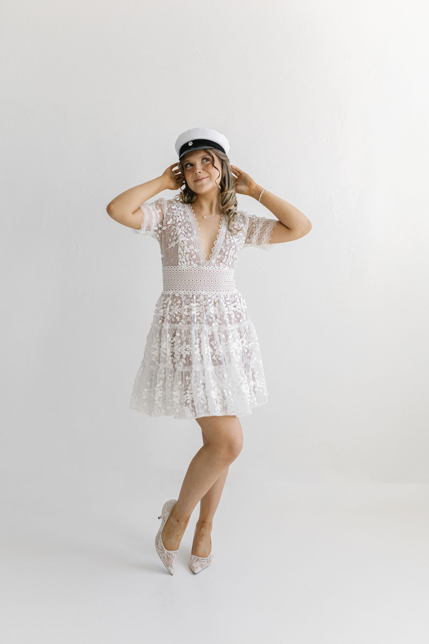Woman in white lace mini dress and white captain hat posing in heels against a white background.