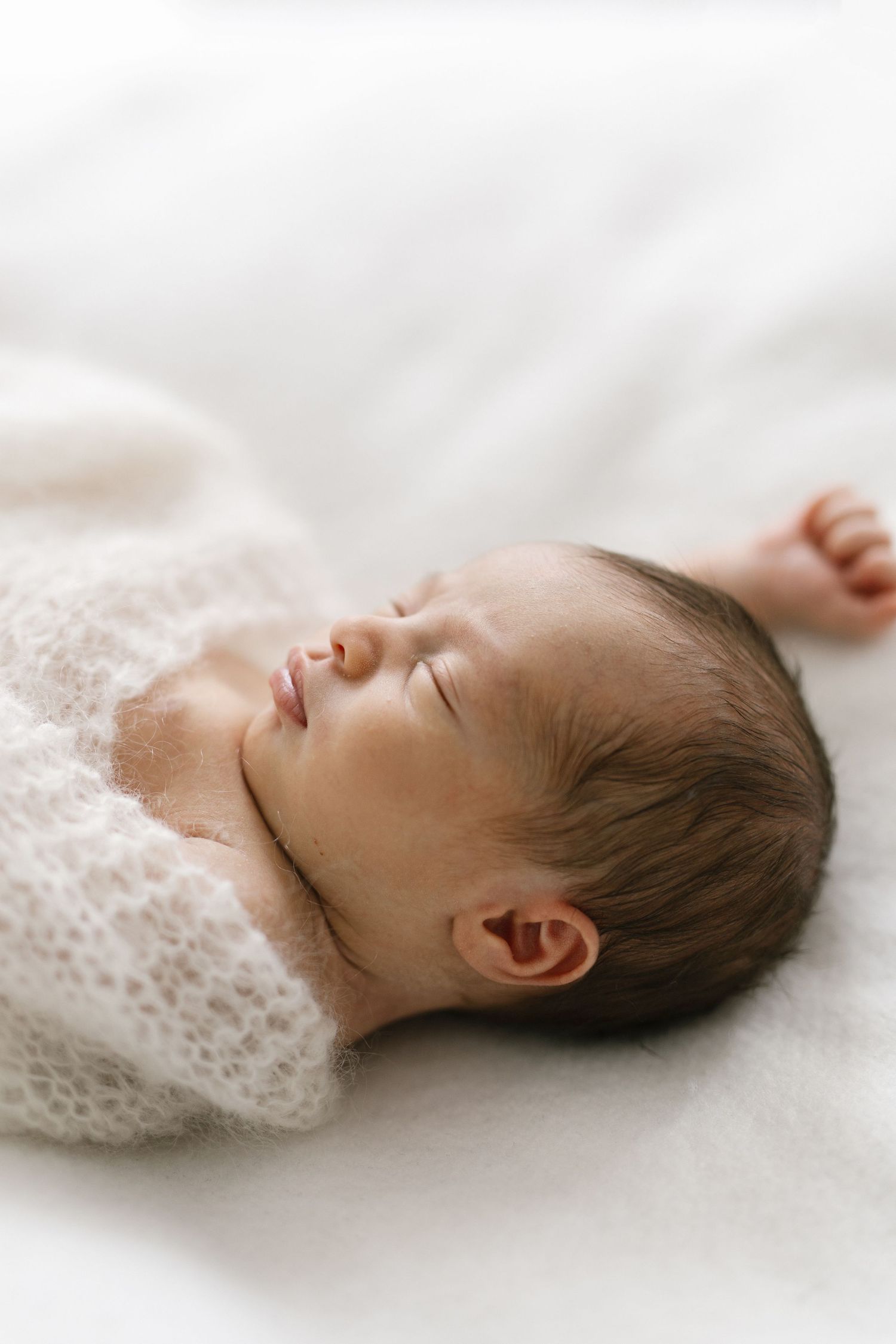 Peaceful newborn baby sleeping wrapped in a soft white knit blanket, with dark hair and tiny clenched fist.