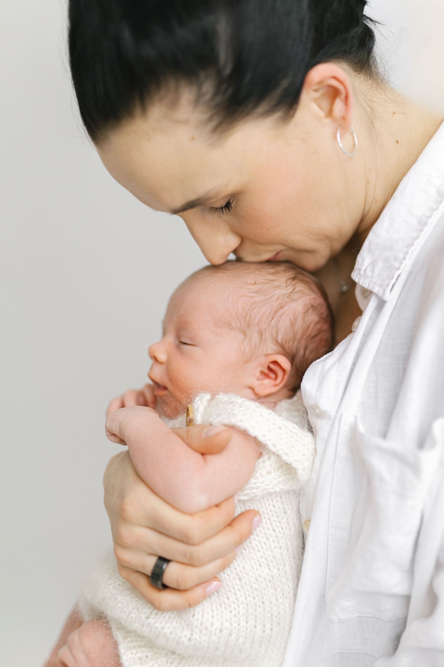 A mother tenderly kisses her sleeping newborn baby dressed in white, captured in a soft, intimate portrait.
