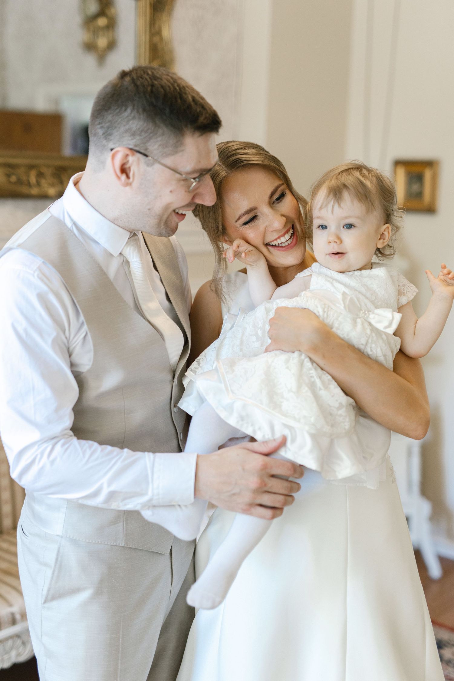 Happy couple in wedding attire smiling at their baby girl in a white dress during a bright indoor ceremony.