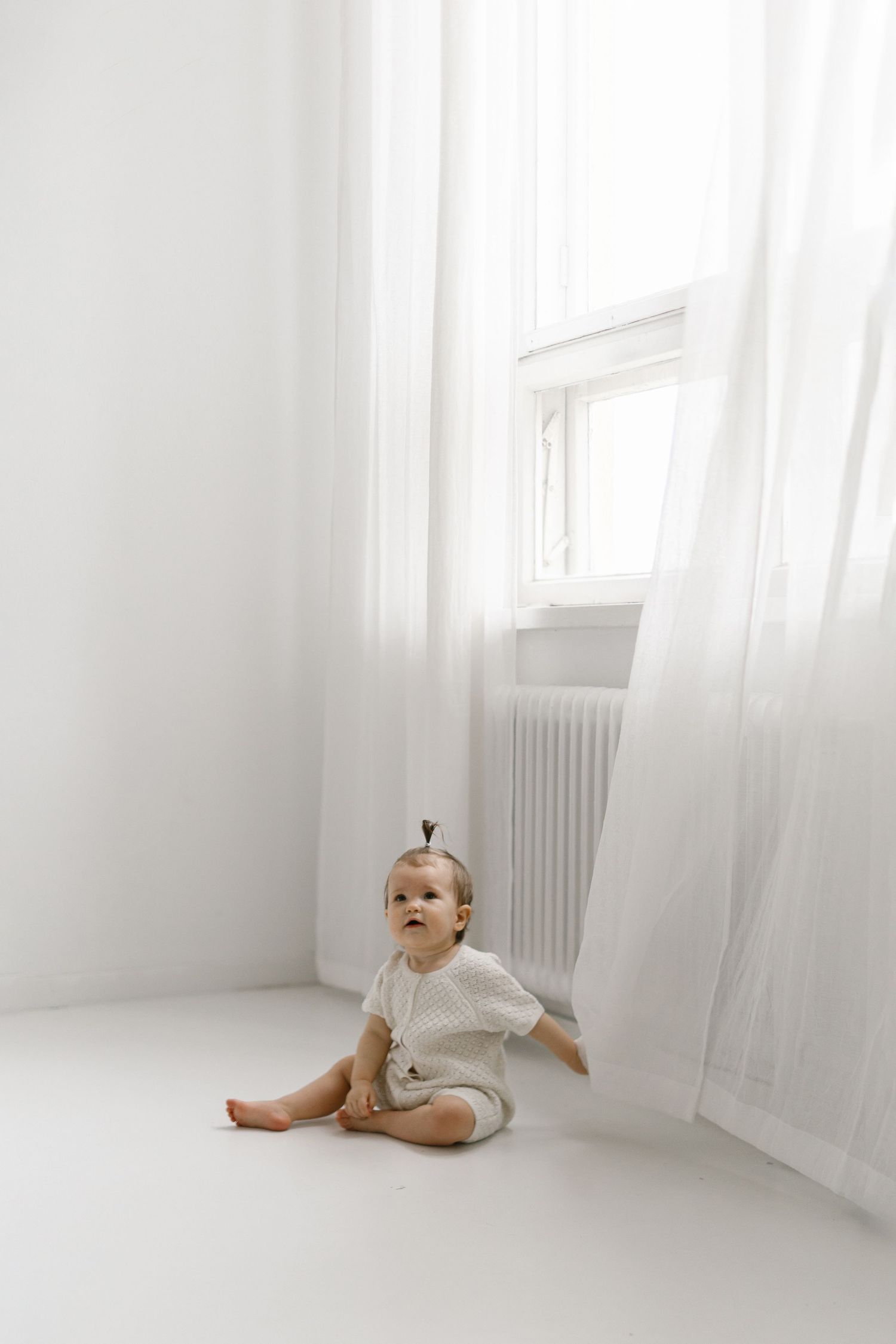 A curious baby in a white romper sits on a bright white floor beside sheer curtains near a window.