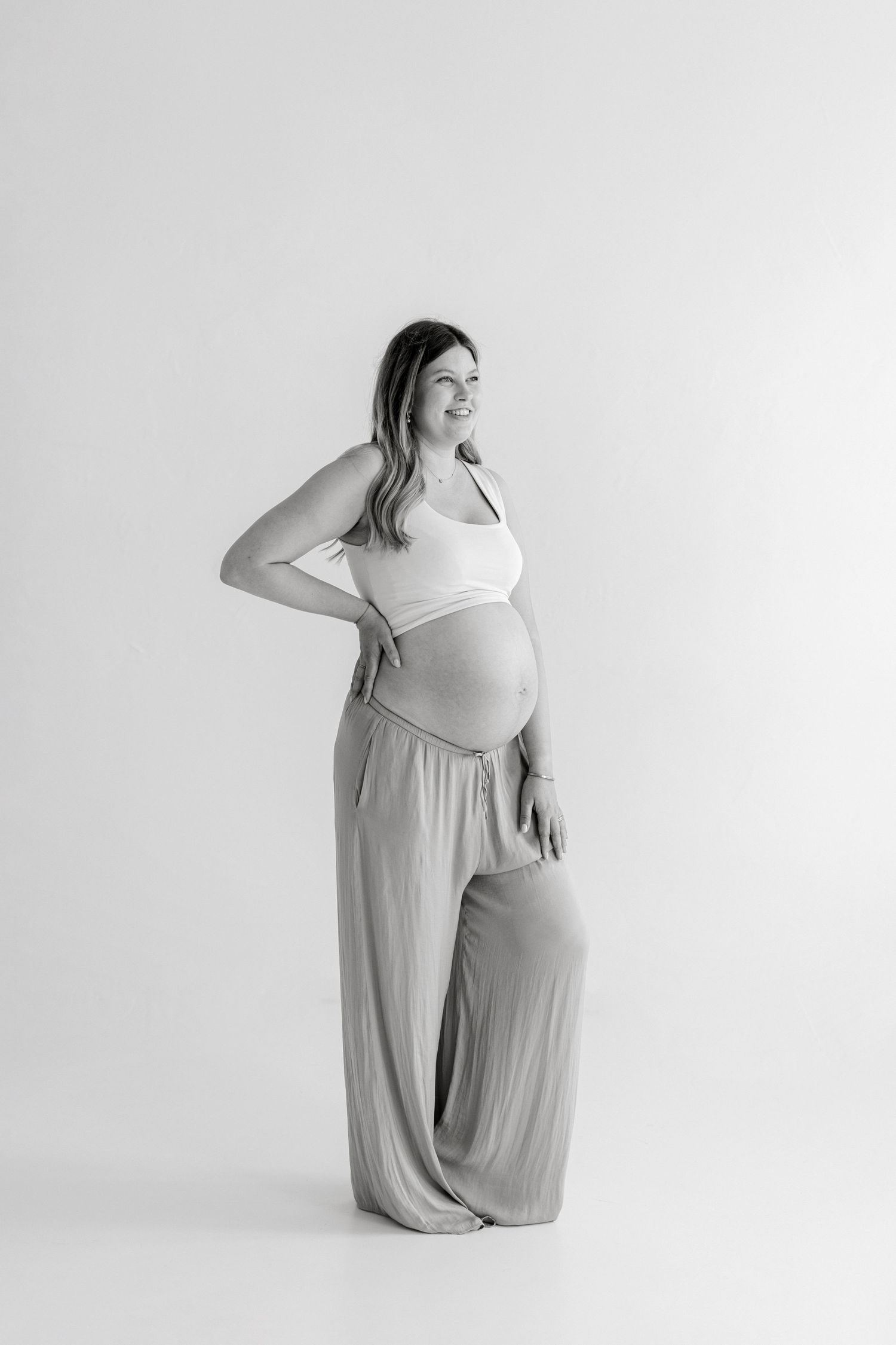Black and white maternity photo of smiling pregnant woman in crop top and wide-leg pants, cradling baby bump in studio.