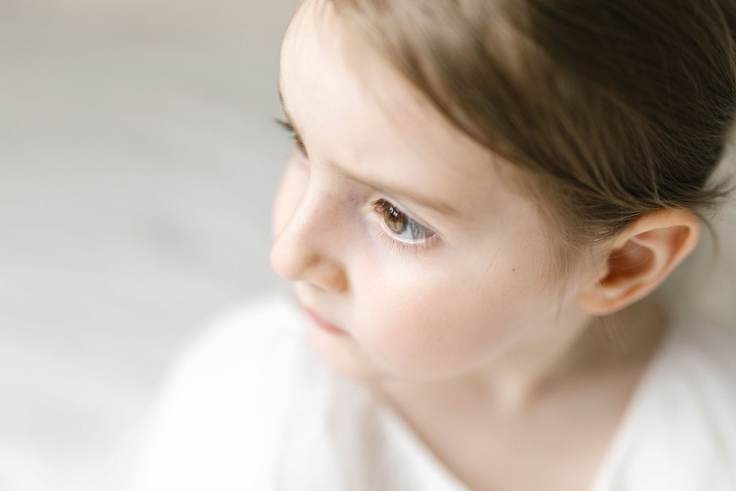 Close-up portrait of a young girl with brown eyes looking up thoughtfully against a soft white background.