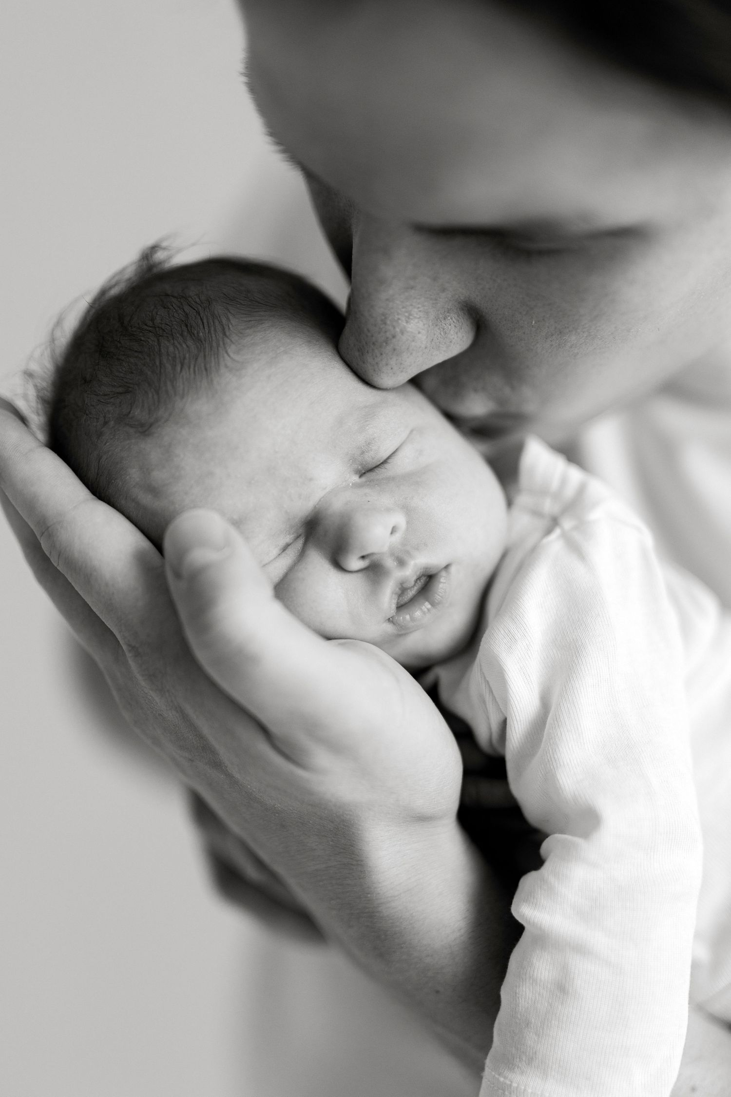 Black and white photo of a mother gently kissing her sleeping newborn baby cradled in her hands.
