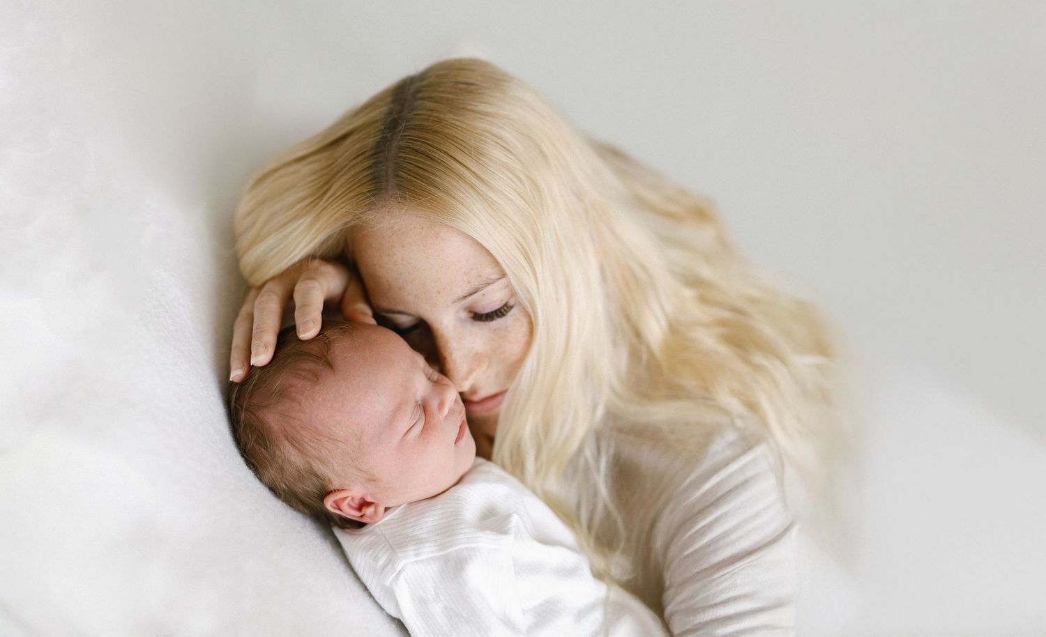 A blonde mother gently kisses her newborn baby wrapped in white, sharing a tender moment against a soft background.
