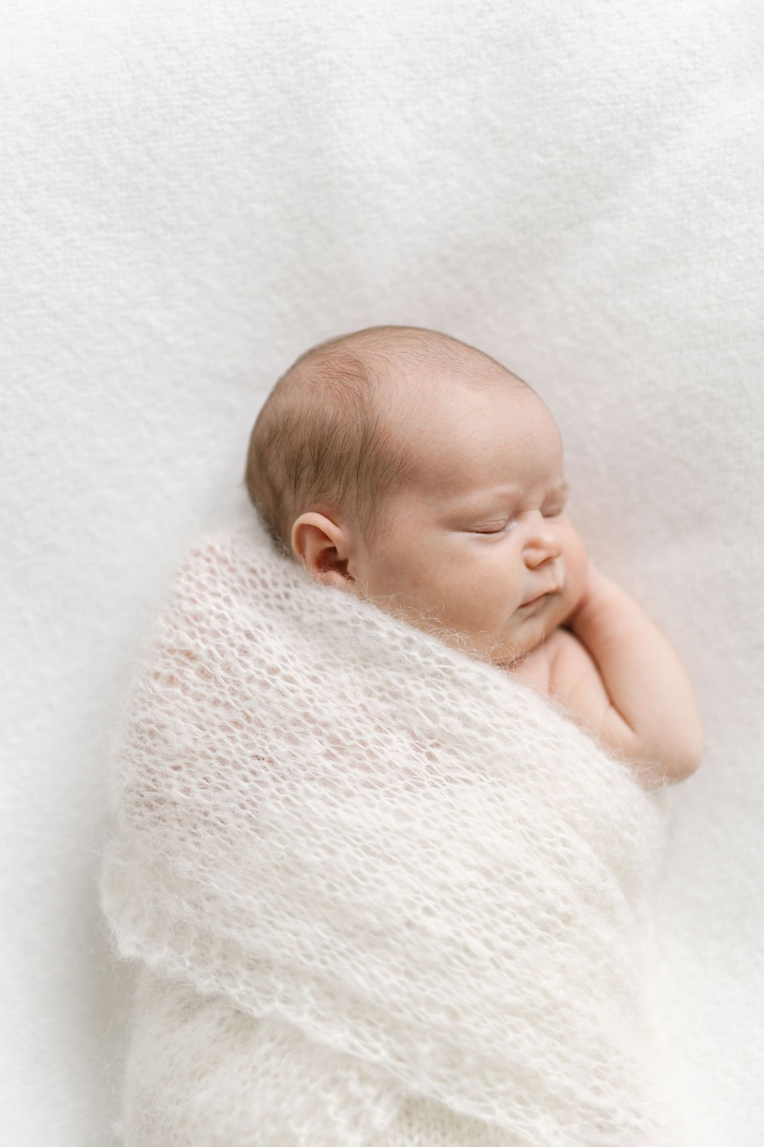 Peaceful newborn baby swaddled in a soft white knit wrap, sleeping on a white fluffy blanket.