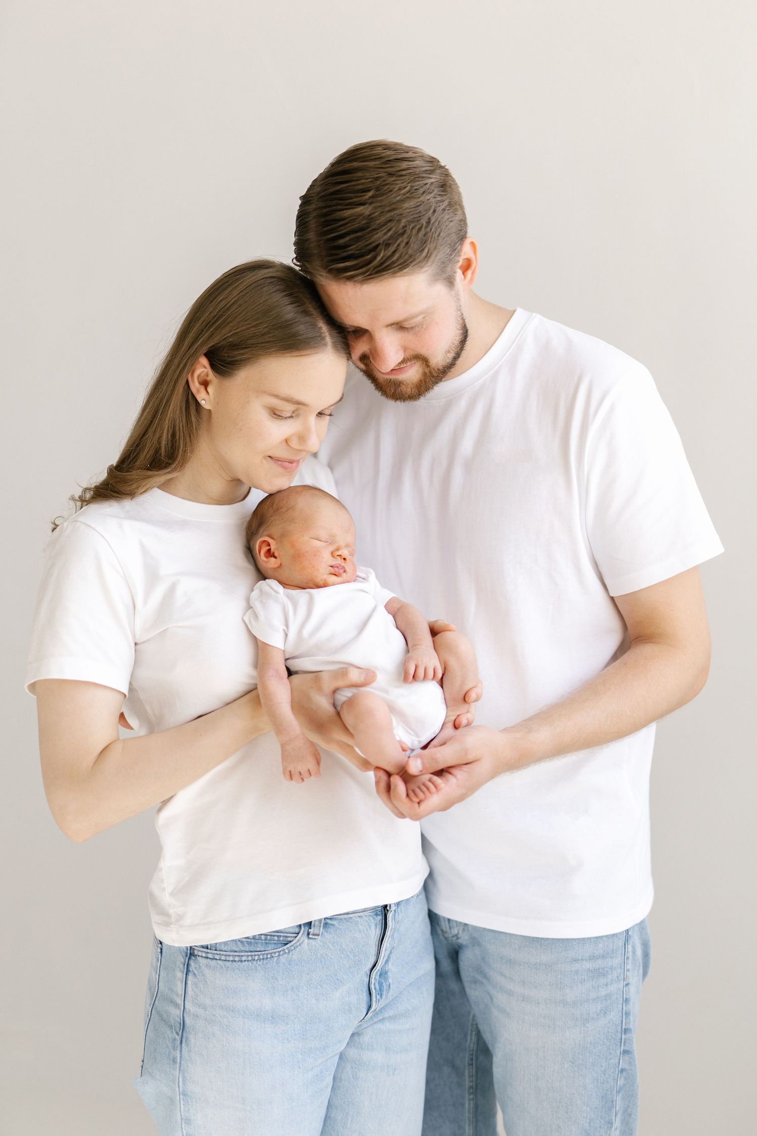 Young parents in white t-shirts and jeans tenderly holding their newborn baby against a light background.