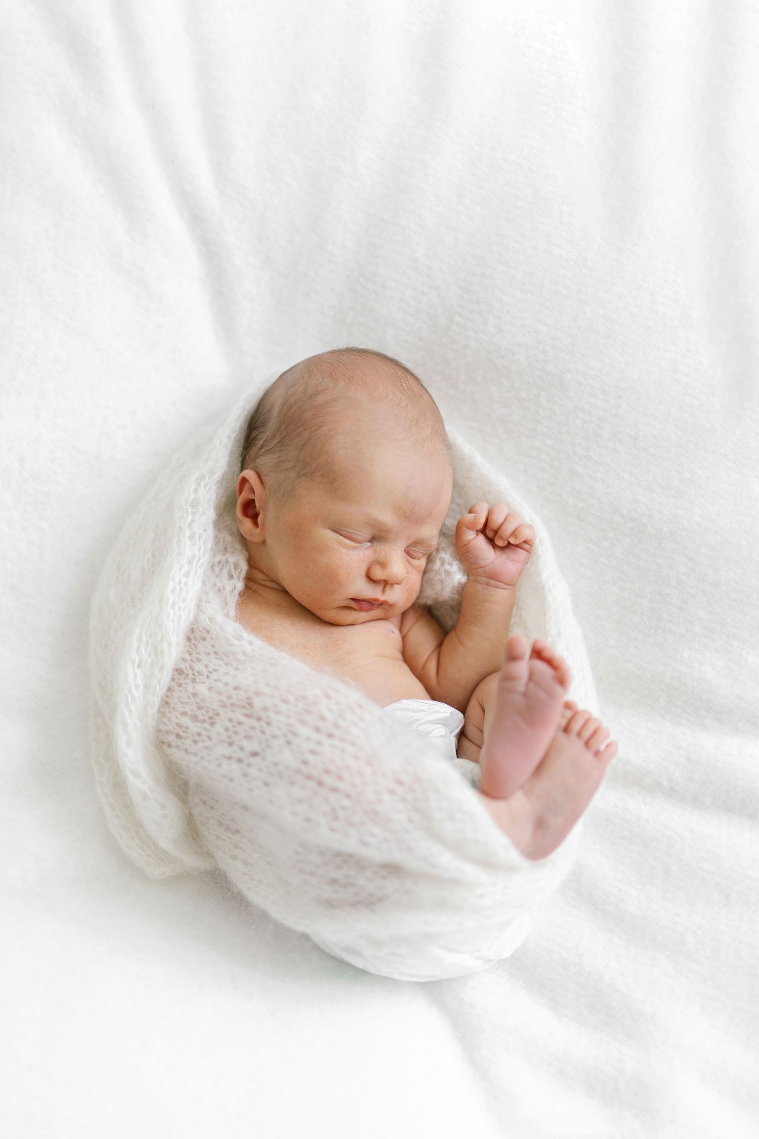 Peaceful newborn baby sleeping swaddled in a soft white wrap on a white blanket.