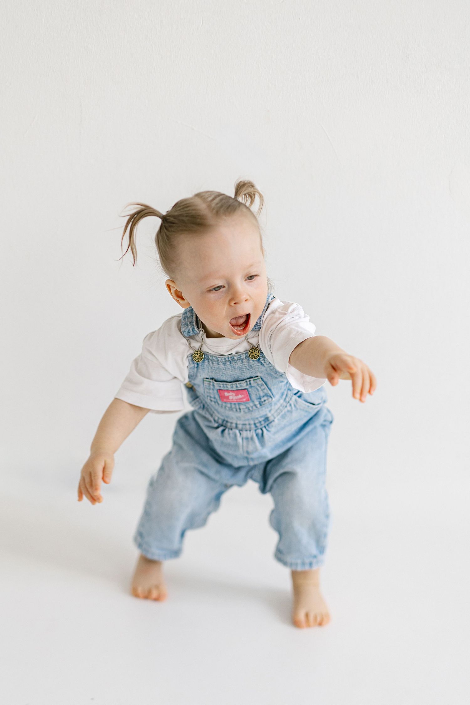 Toddler girl in denim overalls and pigtails walking barefoot with mouth open on white background.