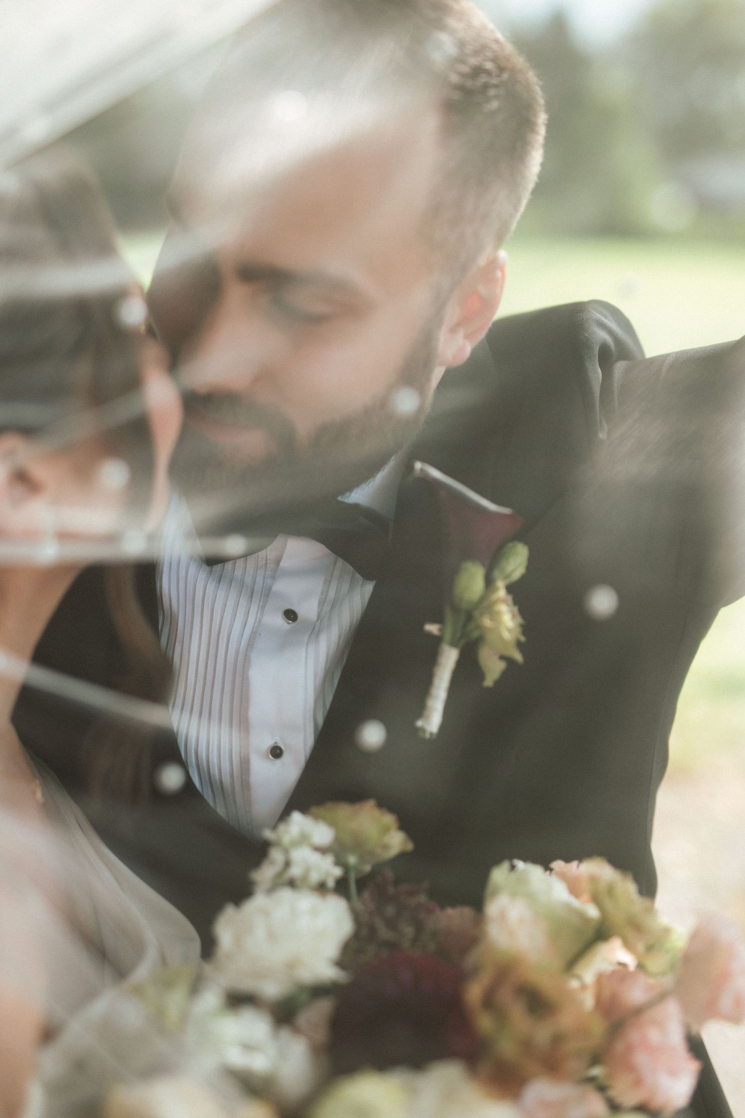 Groom in black tuxedo with floral boutonniere leans toward bride holding bouquet, shot through a veil.