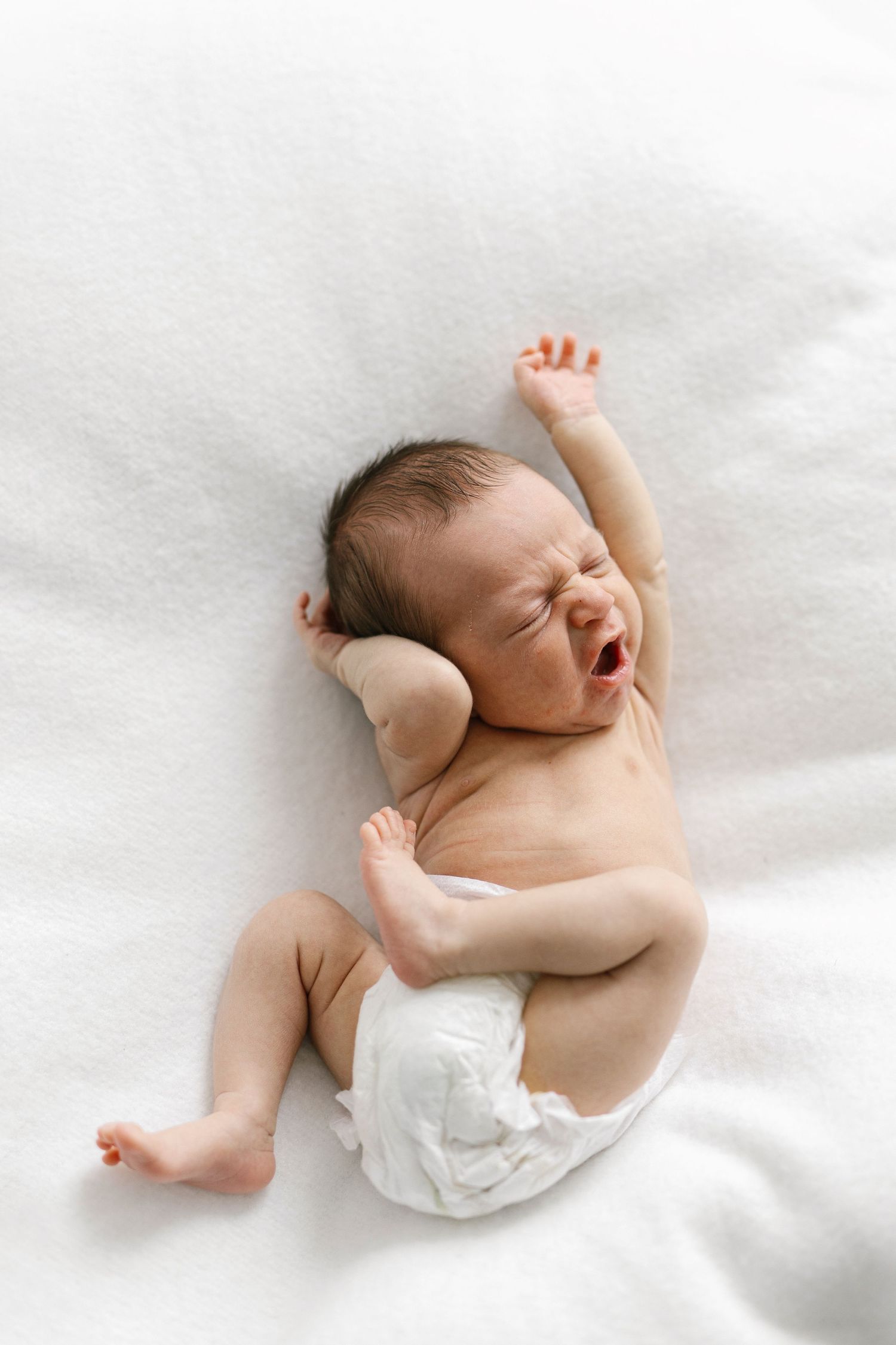 Newborn baby in a diaper lying on white blanket, yawning and stretching arms, capturing a sweet candid moment.