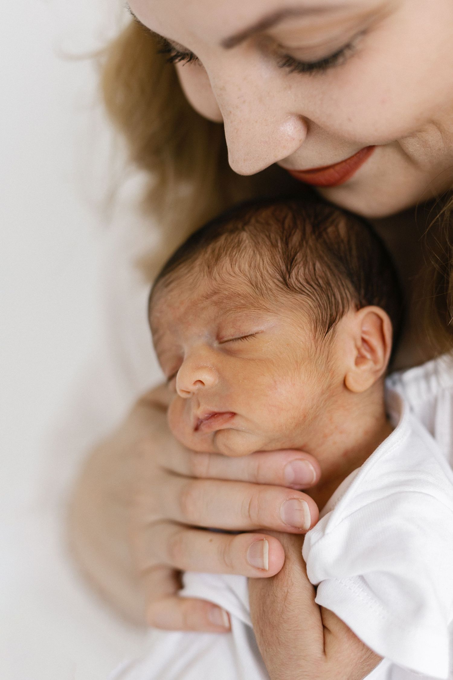 A smiling mother tenderly holds her sleeping newborn baby wrapped in white cloth.