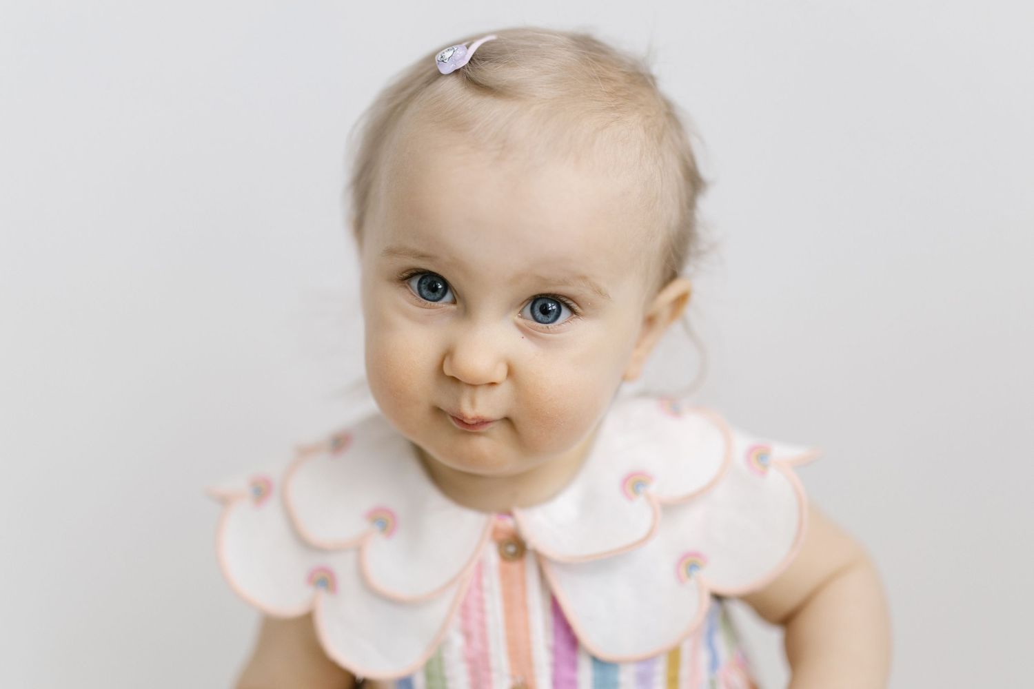 Baby wearing pink scalloped collar and striped dress looks at camera with bright blue eyes against white background.