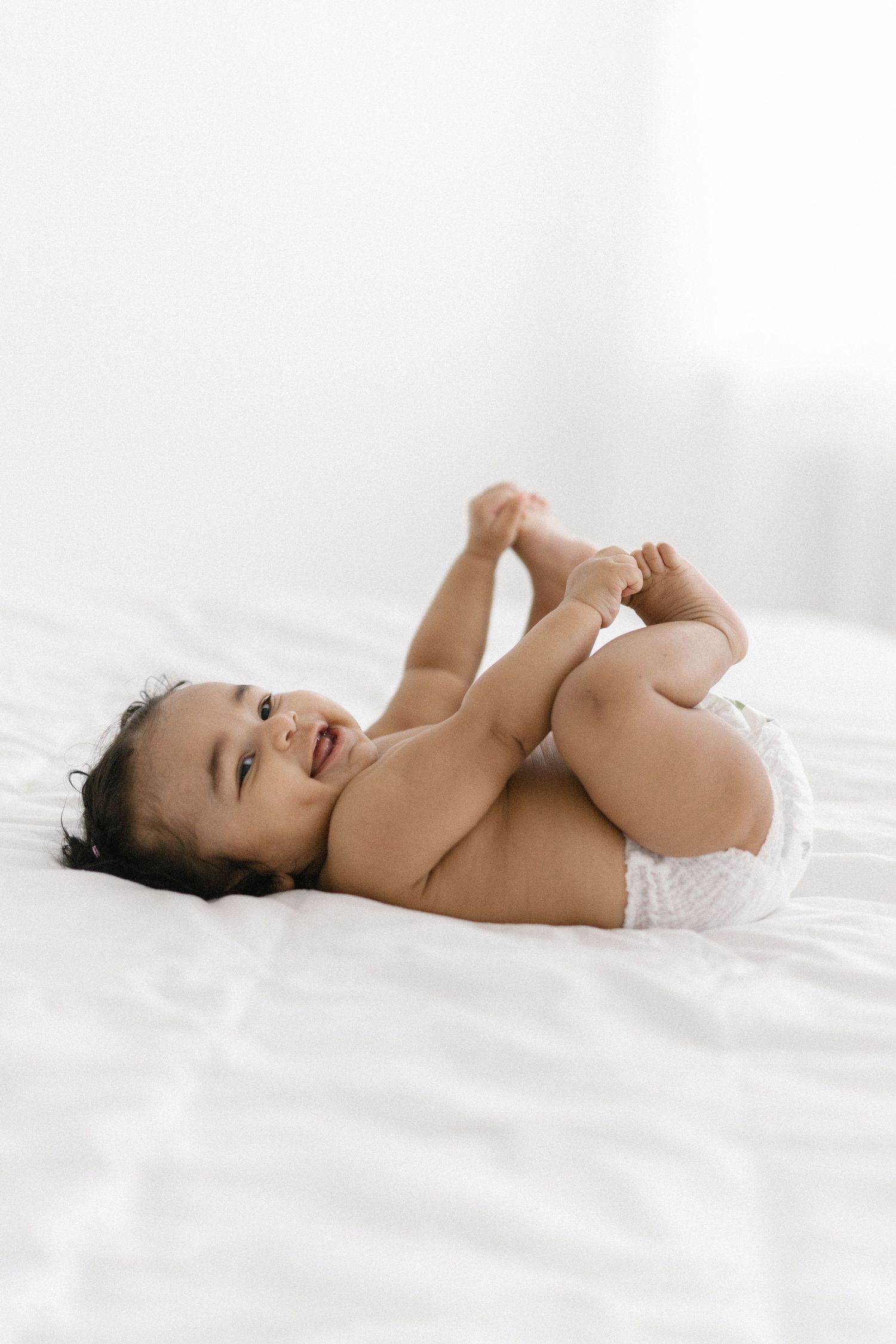 Happy baby lying on white bed in diaper, smiling and holding feet up in adorable playful pose.