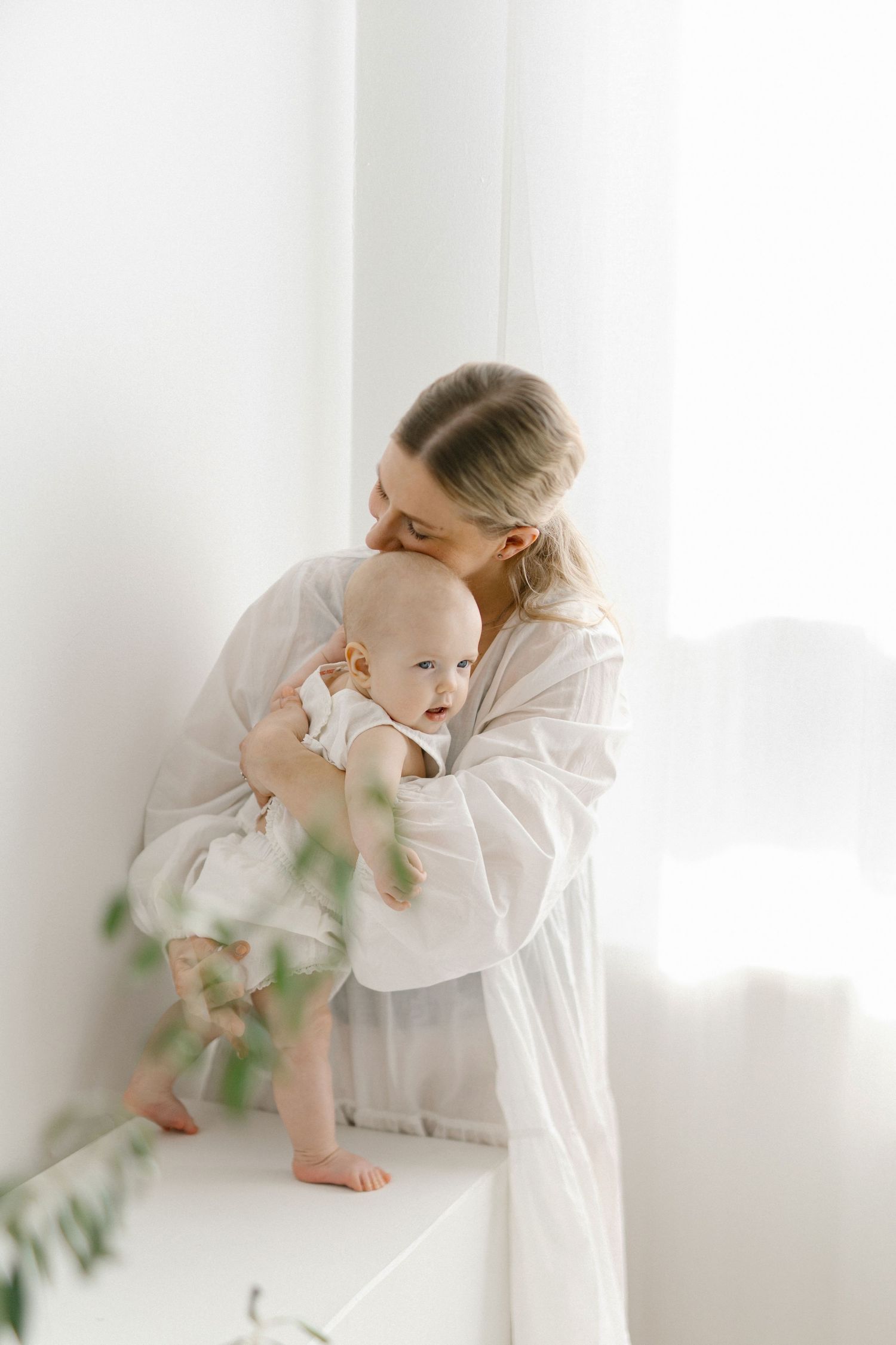 A tender mother in white dress kisses her baby while holding them near a bright window.