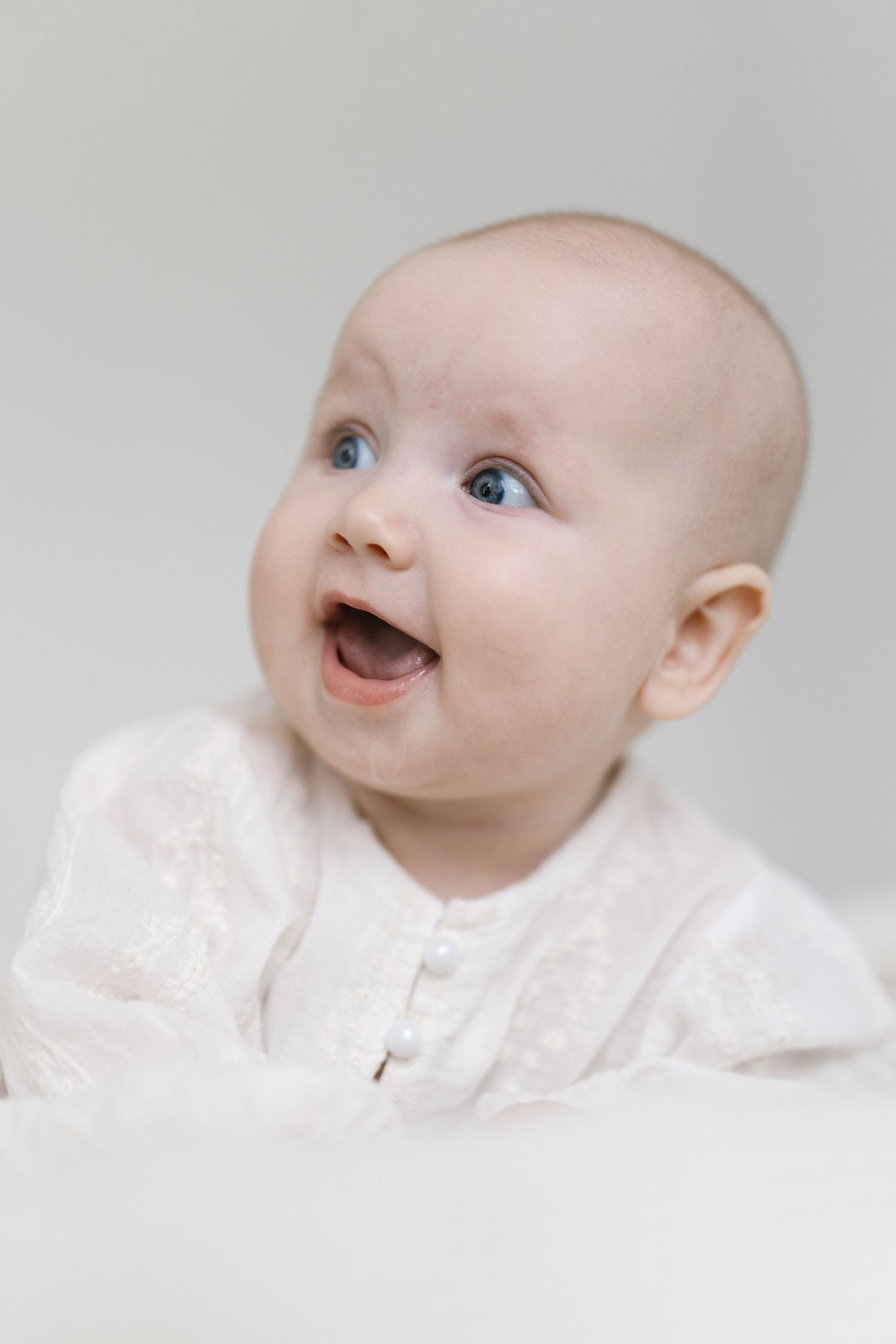 Adorable baby with blue eyes and open mouth smiling, wearing a white lace outfit against a light background.