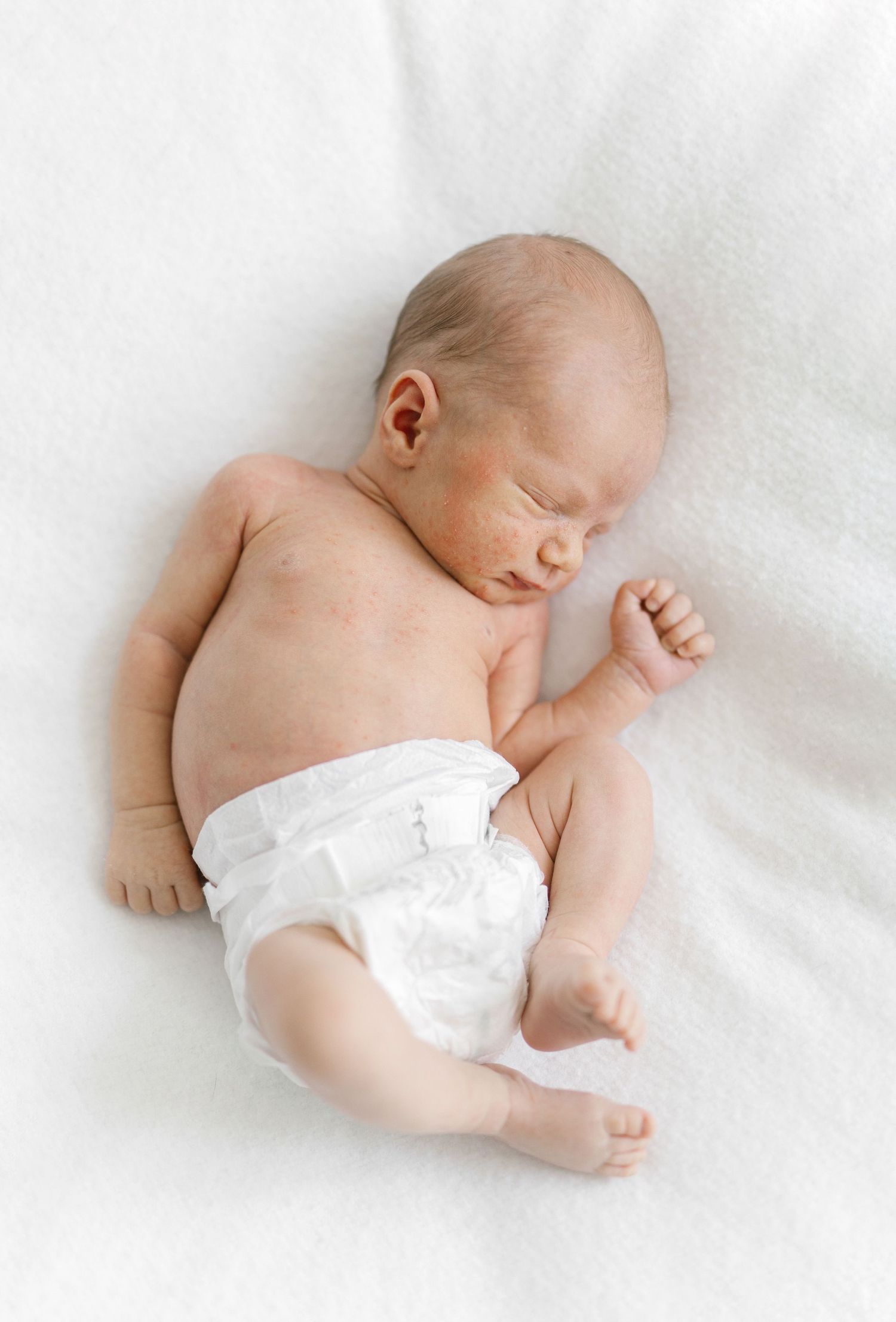 Peaceful newborn baby sleeping on white blanket wearing a white diaper, curled in natural position.
