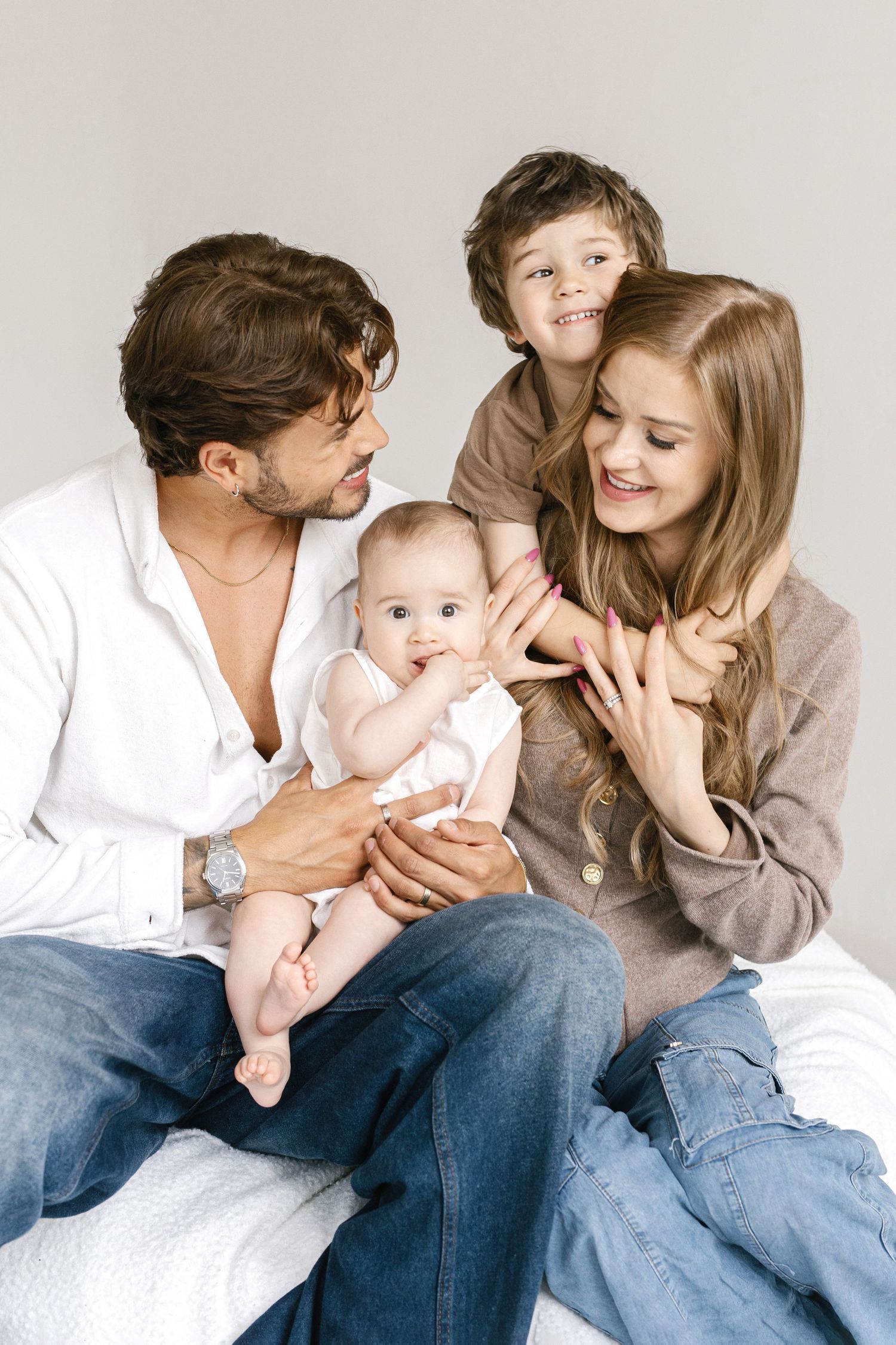 Happy young family with baby and toddler sitting together on bed, smiling and bonding in casual home setting.