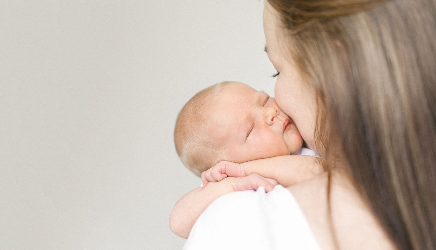 A mother tenderly kisses her sleeping newborn baby resting on her shoulder against a soft white background.