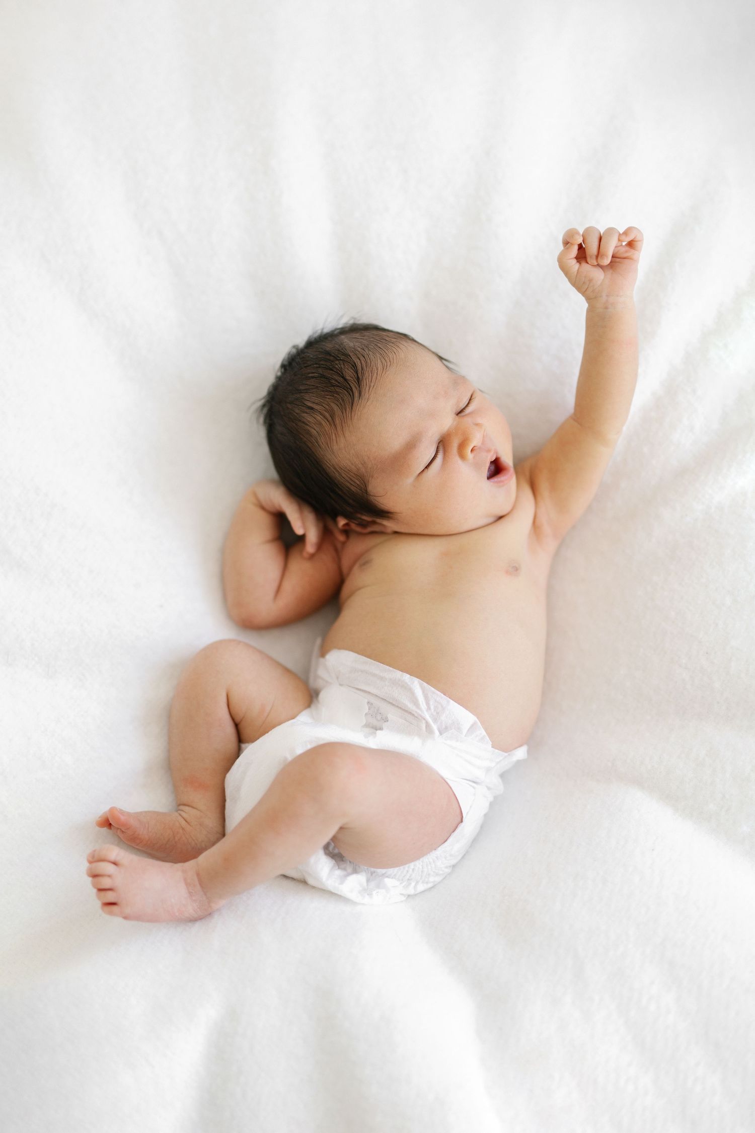 Newborn baby in white diaper lying on soft white blanket, stretching one arm up with mouth open.