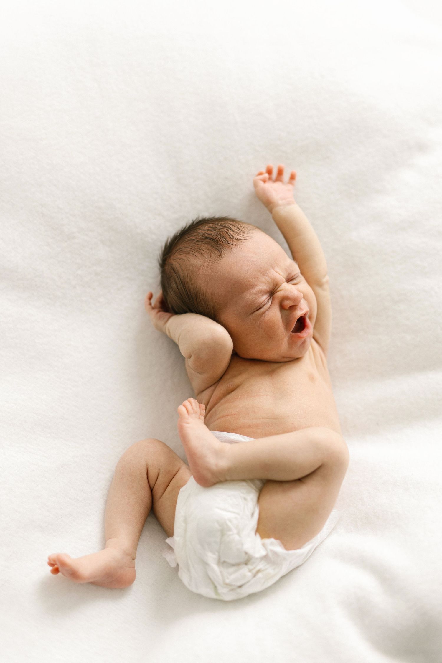 Newborn baby in a diaper lying on white blanket, yawning and stretching arms, peaceful infant portrait.