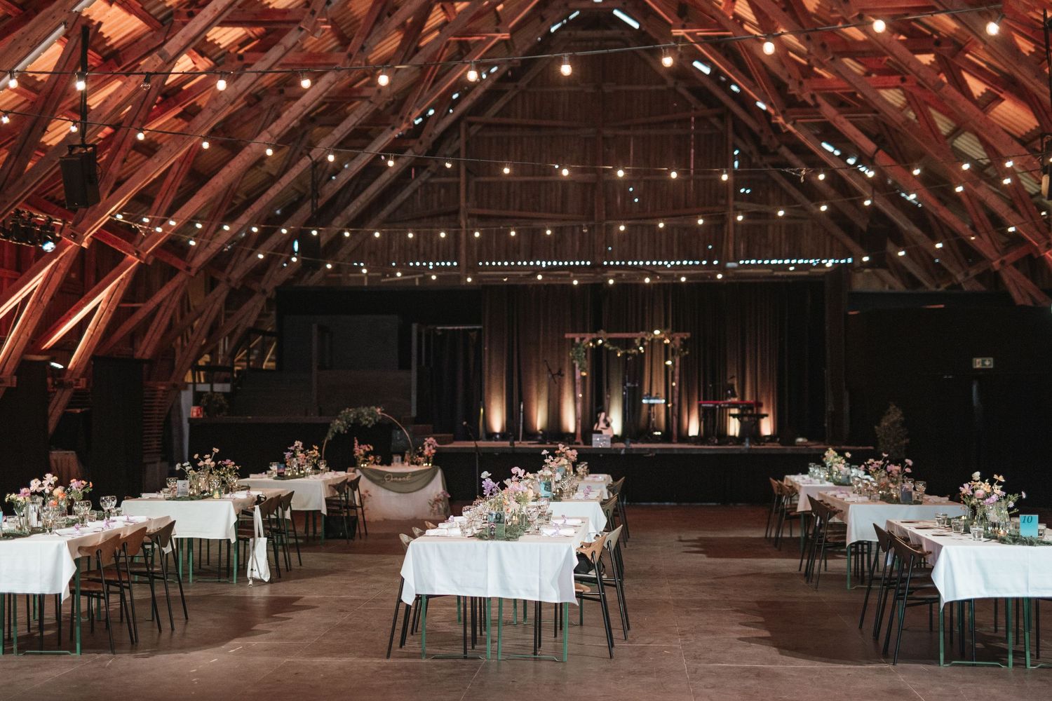 Rustic barn wedding reception with string lights, white tablecloths, floral centerpieces, and warm wood beam ceiling.