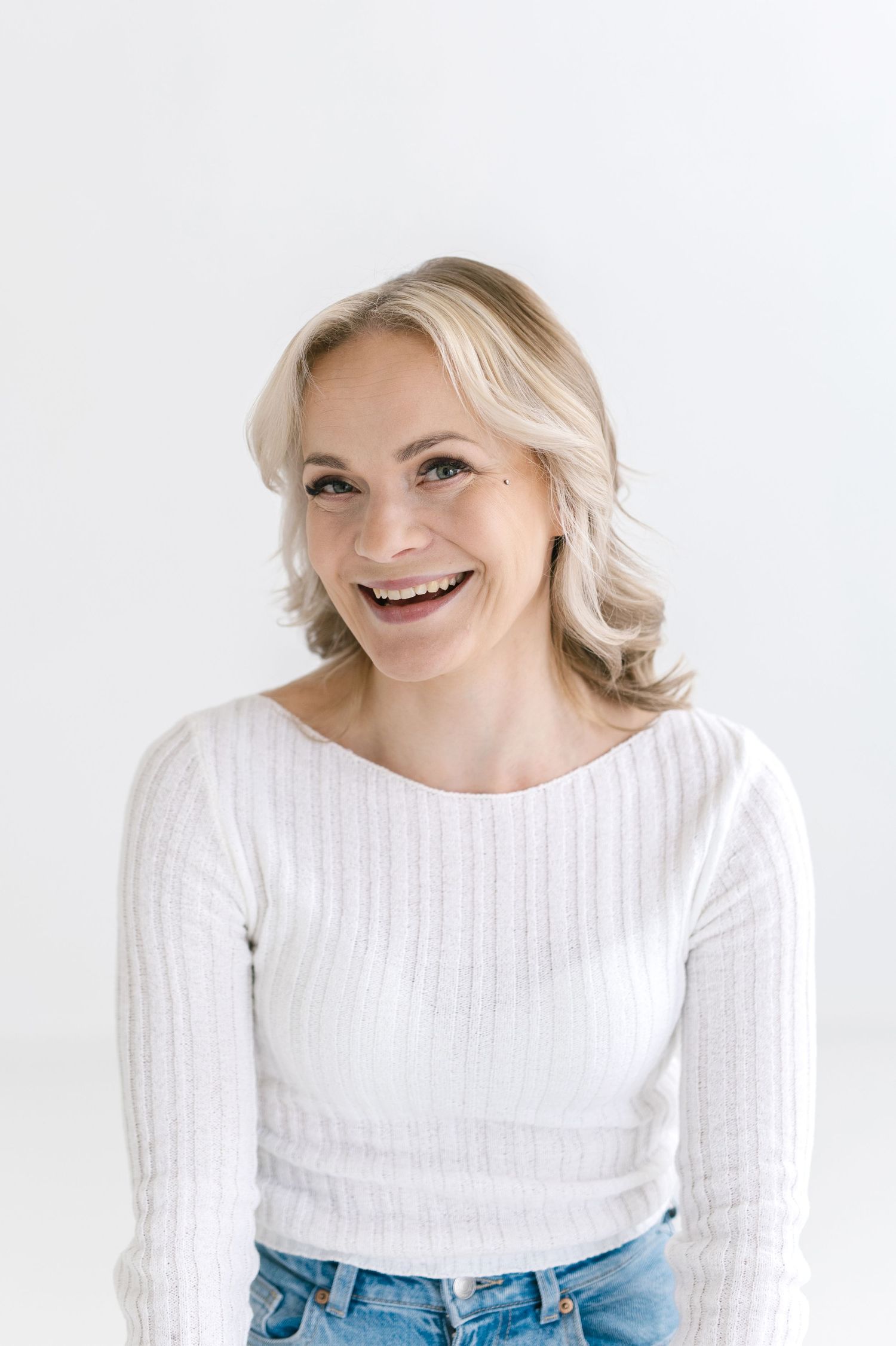 A person wearing a white knit sweater and jeans poses with a bright smile against a light background.