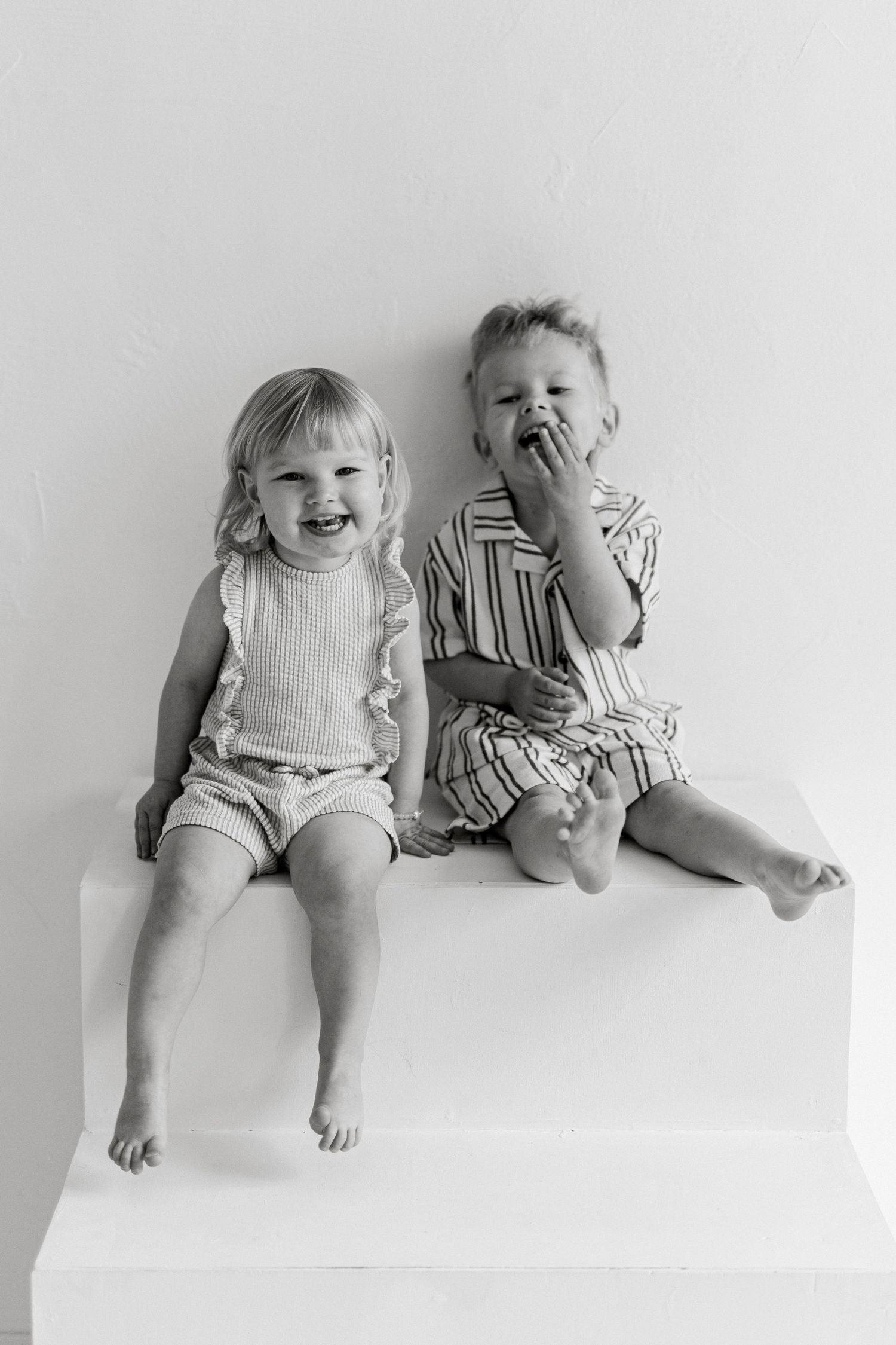 Two laughing toddlers sit barefoot side by side on white steps in a black and white studio portrait of playful siblings.