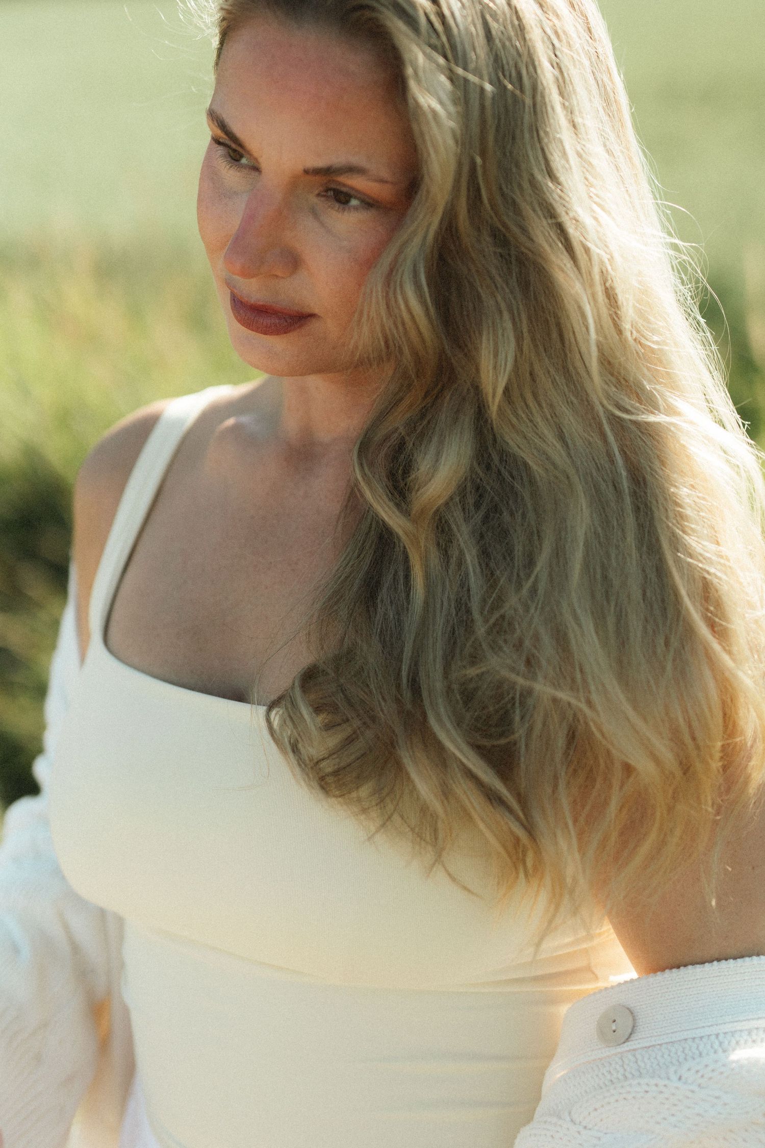 A young blonde woman with wavy hair wearing a white top poses outdoors in soft golden sunlight.