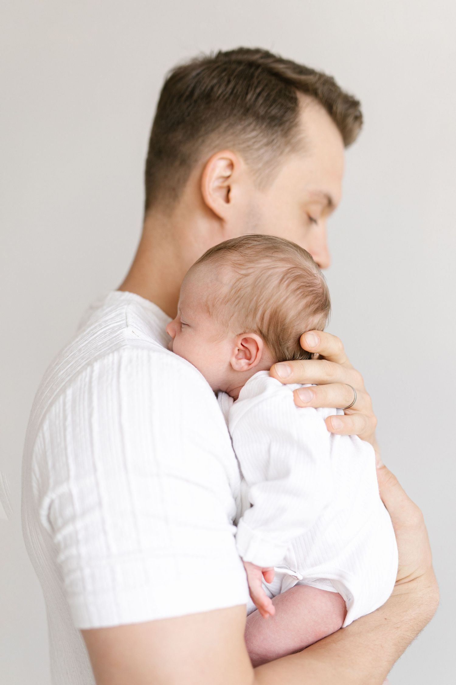 Father in white shirt tenderly holds sleeping newborn baby close to his chest.